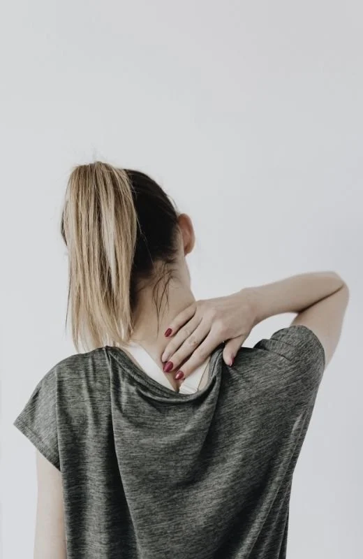 A woman with blonde hair tied in a ponytail, wearing a grey shirt, is holding her neck with her right hand against a plain white background.