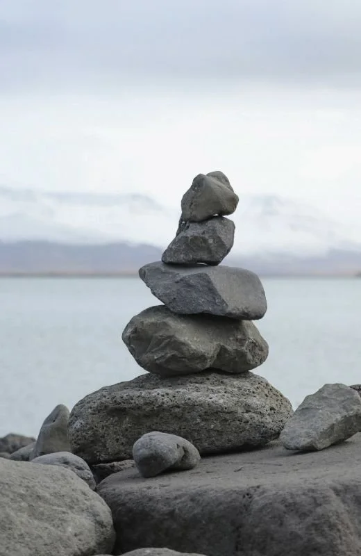 A stack of five rocks balanced on top of each other on the beach with a body of water and cloudy sky in the background.