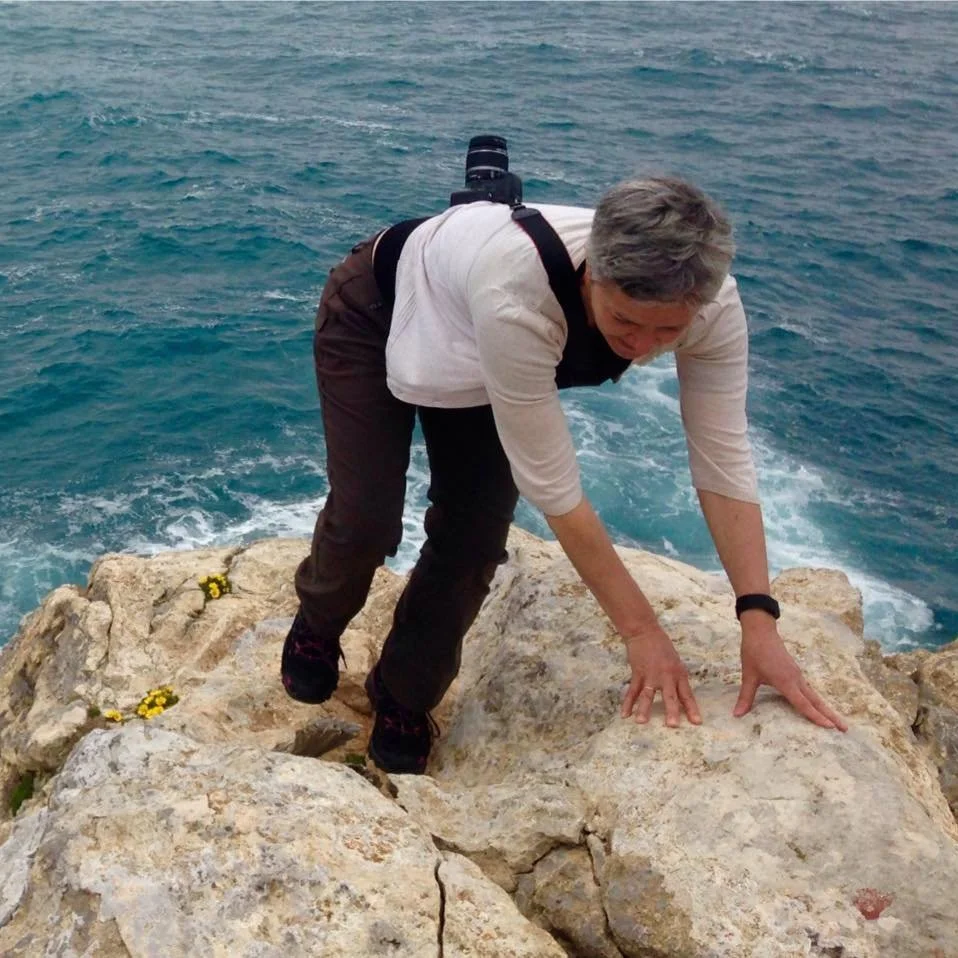 A person with gray hair, wearing a white long sleeve shirt and black pants, is climbing on large rocks by the ocean. They have a camera strapped to their back and are looking downward while gripping the rocks with both hands.