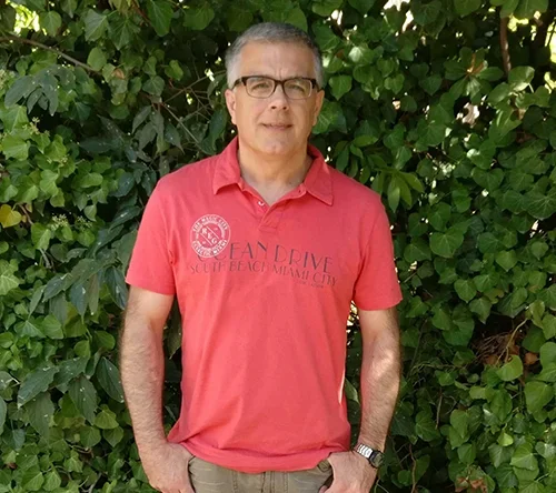 Middle-aged man standing outdoors in front of green foliage, wearing glasses, a red polo shirt, and beige pants.