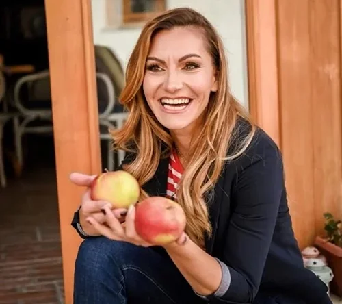 A woman with long blonde hair smiling and holding two apples, one yellow and one red, indoors with wooden walls.