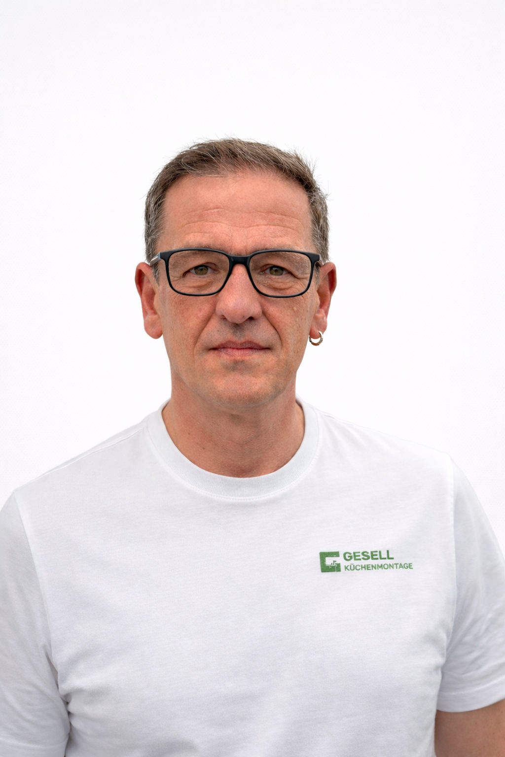 Freundlich, professionell lächelnder Mann (Geschäftsführer) mit Brille, Ohrpiercing, weißem T-Shirt mit Firmen-Logo, grau-weißem Hintergrund.