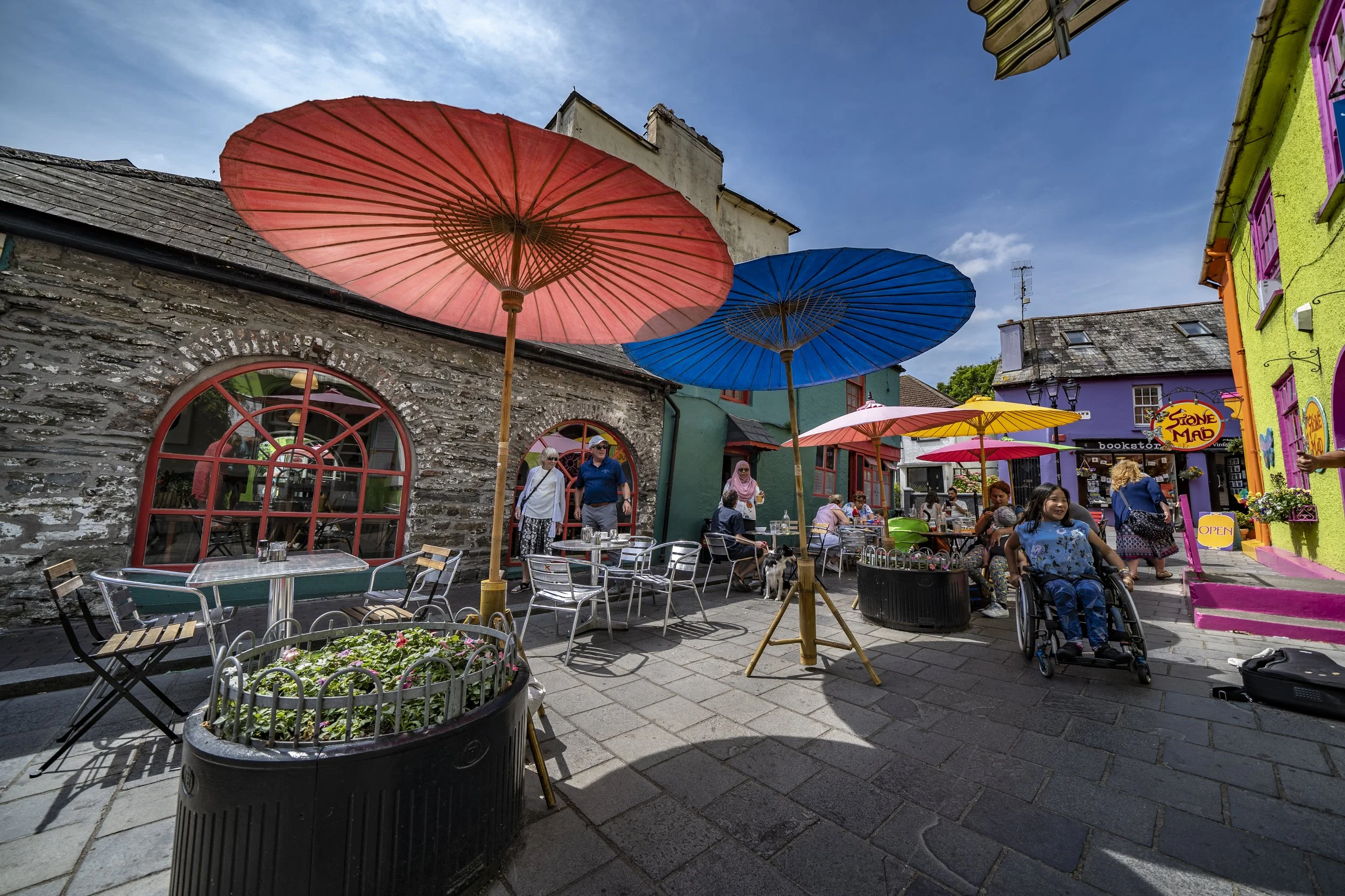 Outdoor cafe with colorful umbrellas and chairs, surrounded by vibrant buildings, some people sitting and walking, and a woman in a wheelchair, on a bright sunny day.