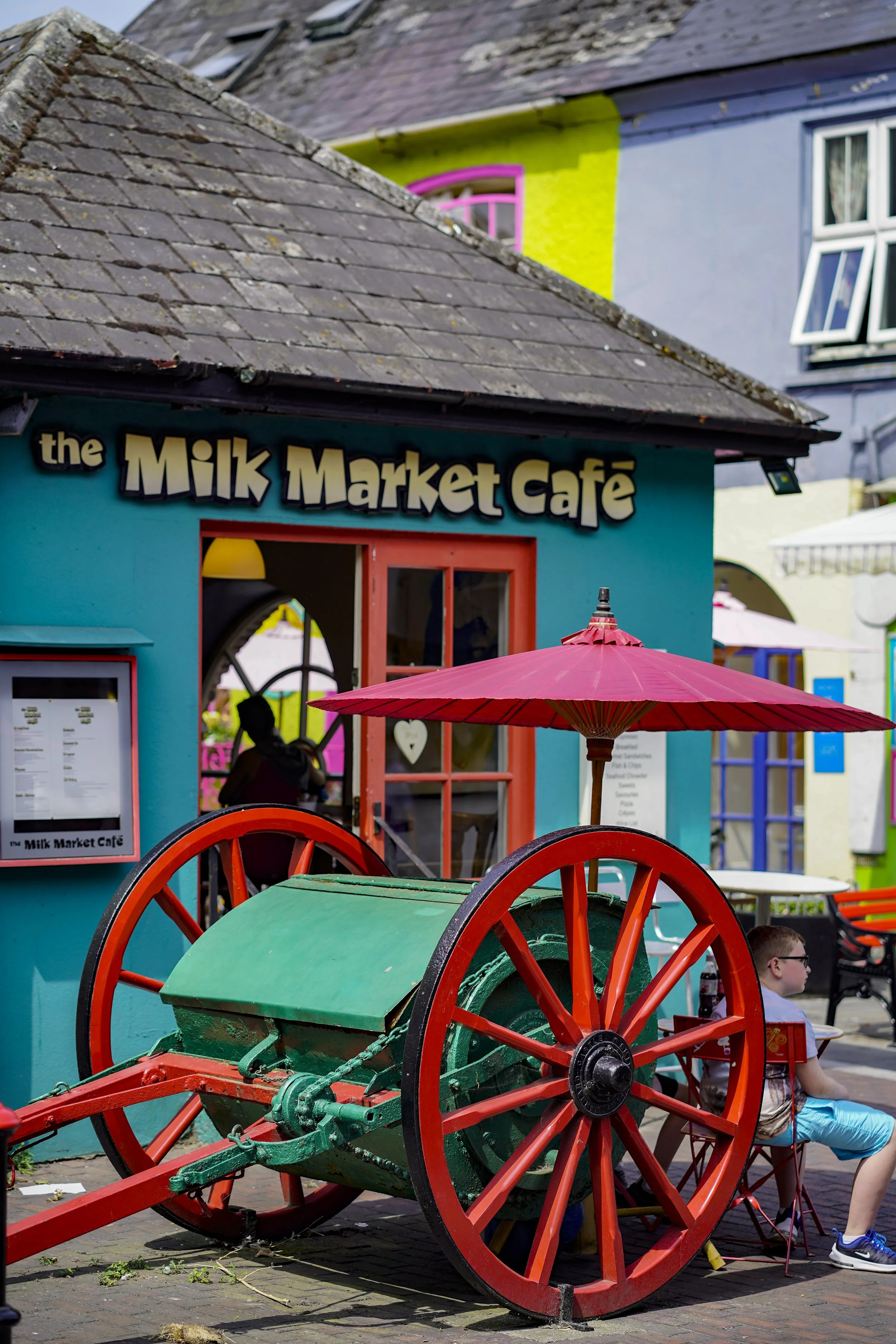 Colorful building with a sign that reads 'the Milk Market Café', a red umbrella, an old green and red cart, and a young boy sitting outside.