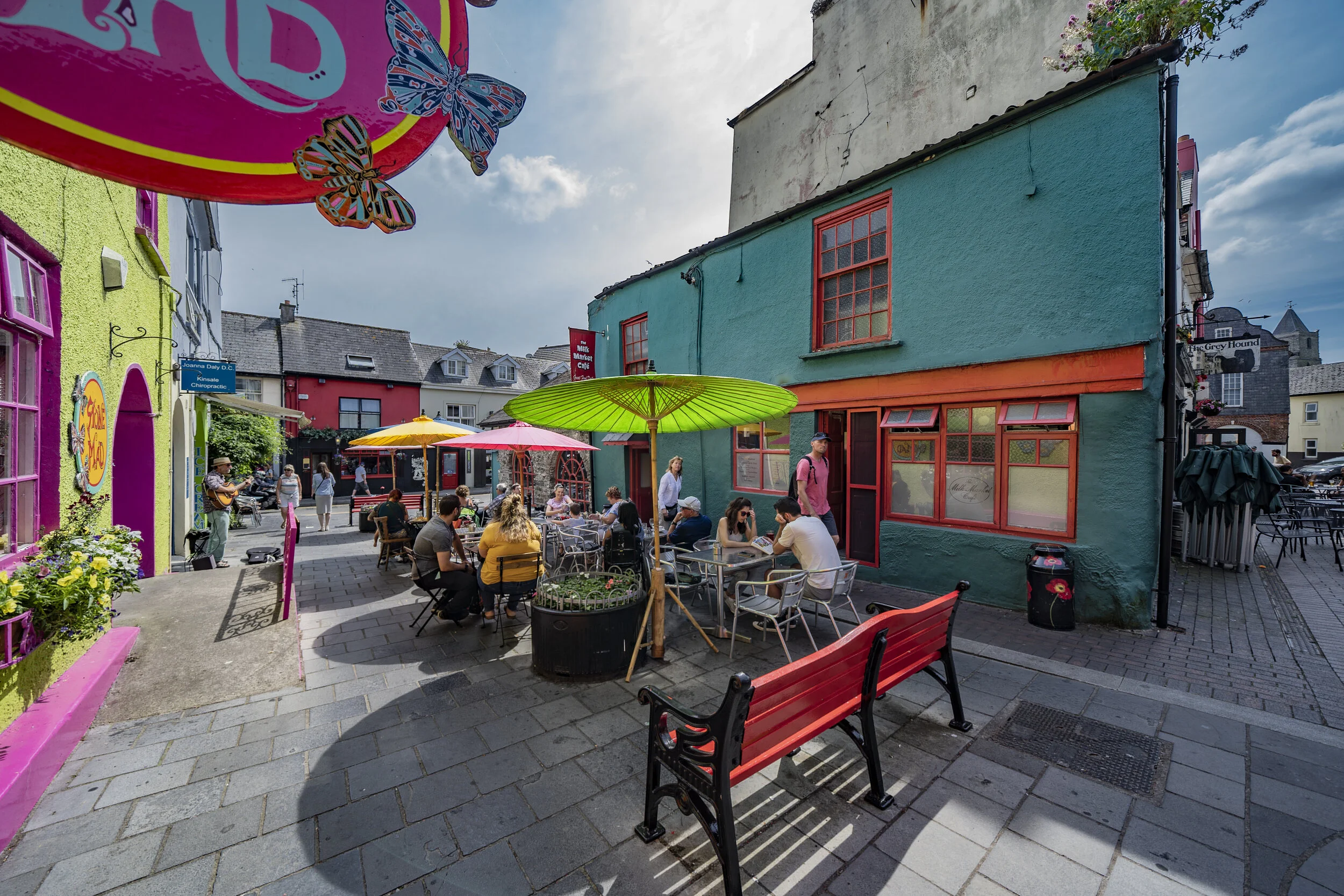 Outdoor seating area in a colorful urban street with people dining under umbrellas, surrounding vibrant buildings with painted facades, and a mural with butterflies visible in the background.