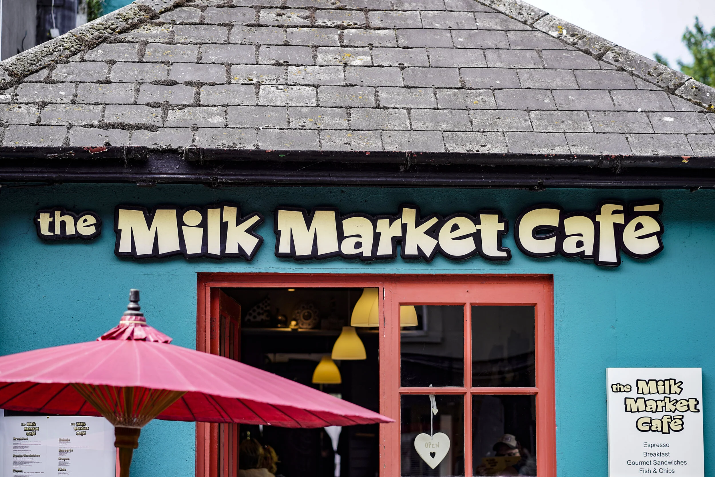 Exterior of the Milk Market Café with a bright blue wall, a red window frame, a pink patio umbrella, and a sign displaying the café's menu.