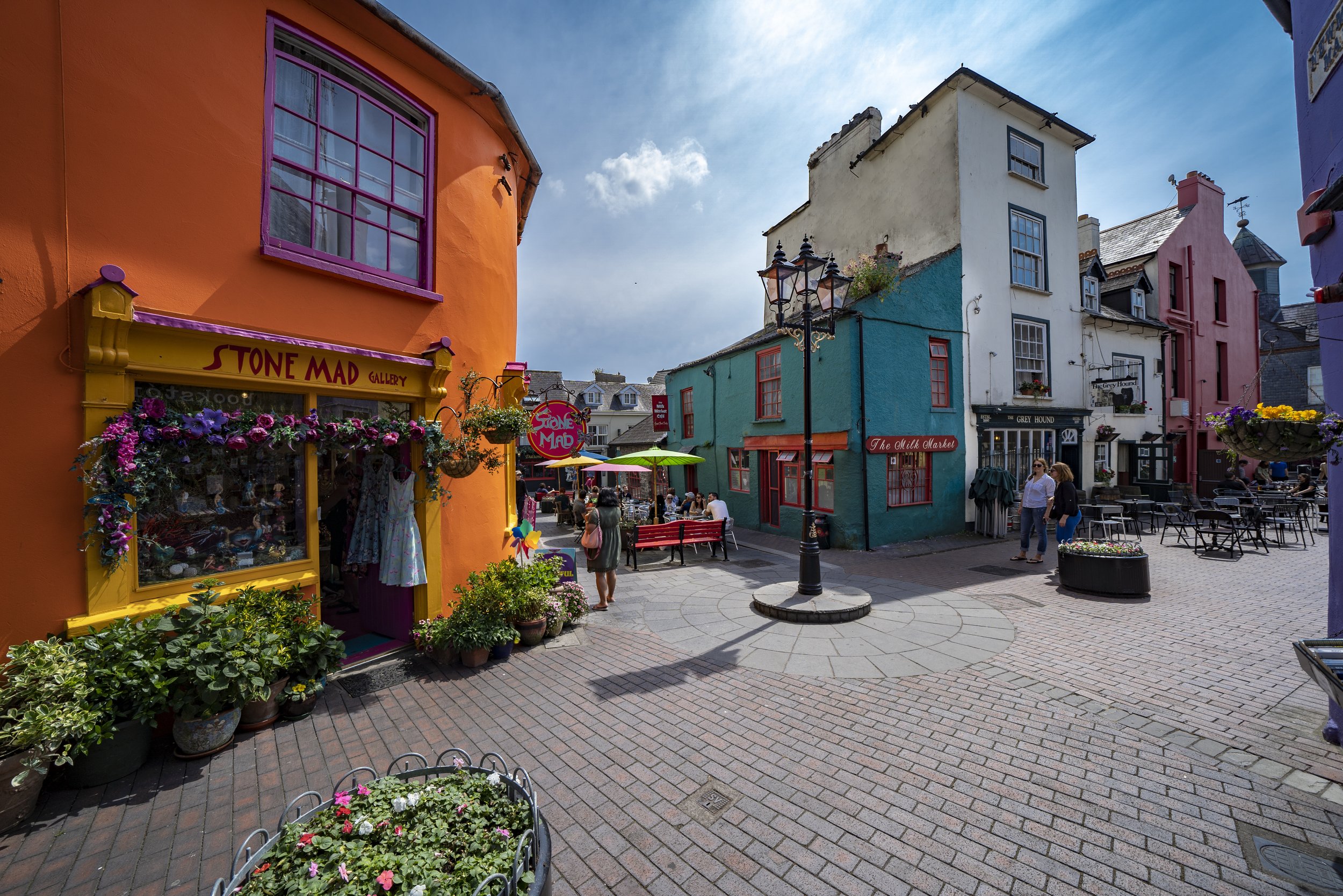 Colorful buildings with shopfronts and outdoor seating on a sunny street, with people walking and chatting.