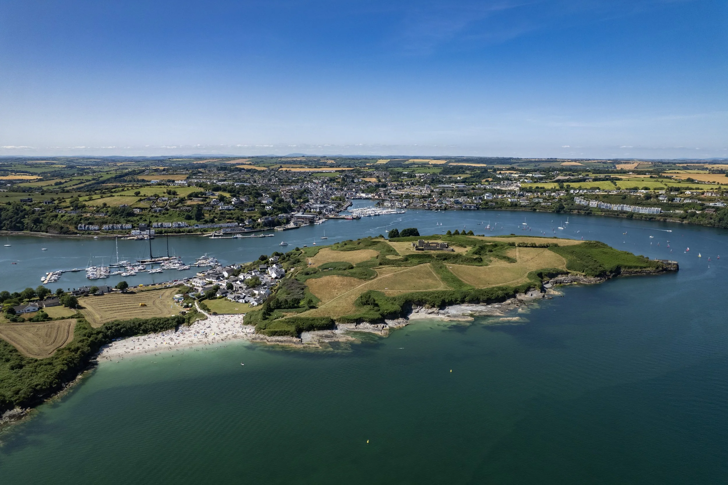 An aerial view of a coastal town featuring a peninsula with green fields, a beach, and a harbor filled with boats. The town extends along the water with residential and commercial buildings, and there are sailboats on the water under a clear blue sky