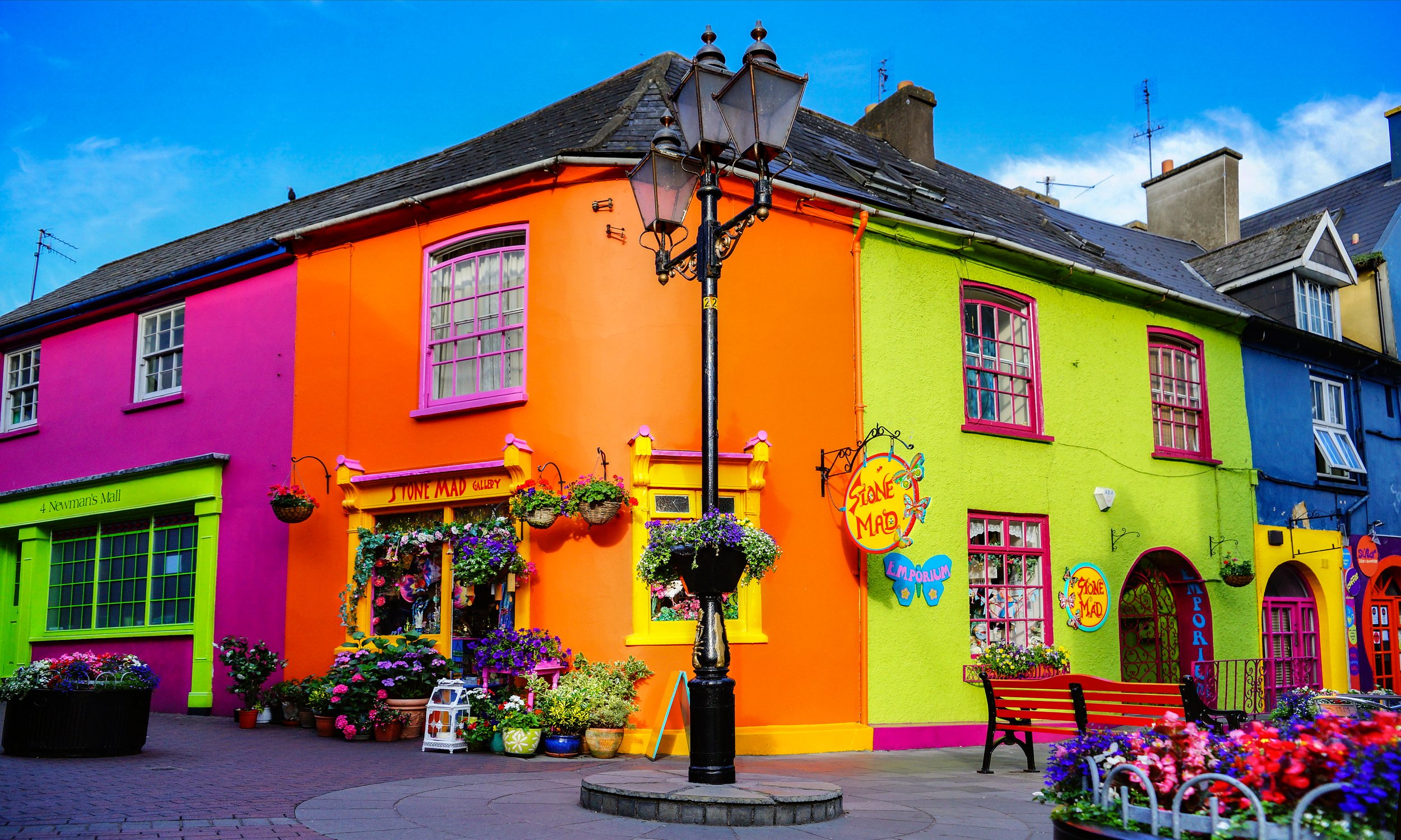 Colorful row of buildings painted in pink, orange, green, and blue with flower baskets, signs, and benches in front of them, on a city street with a black lamp post in the center.