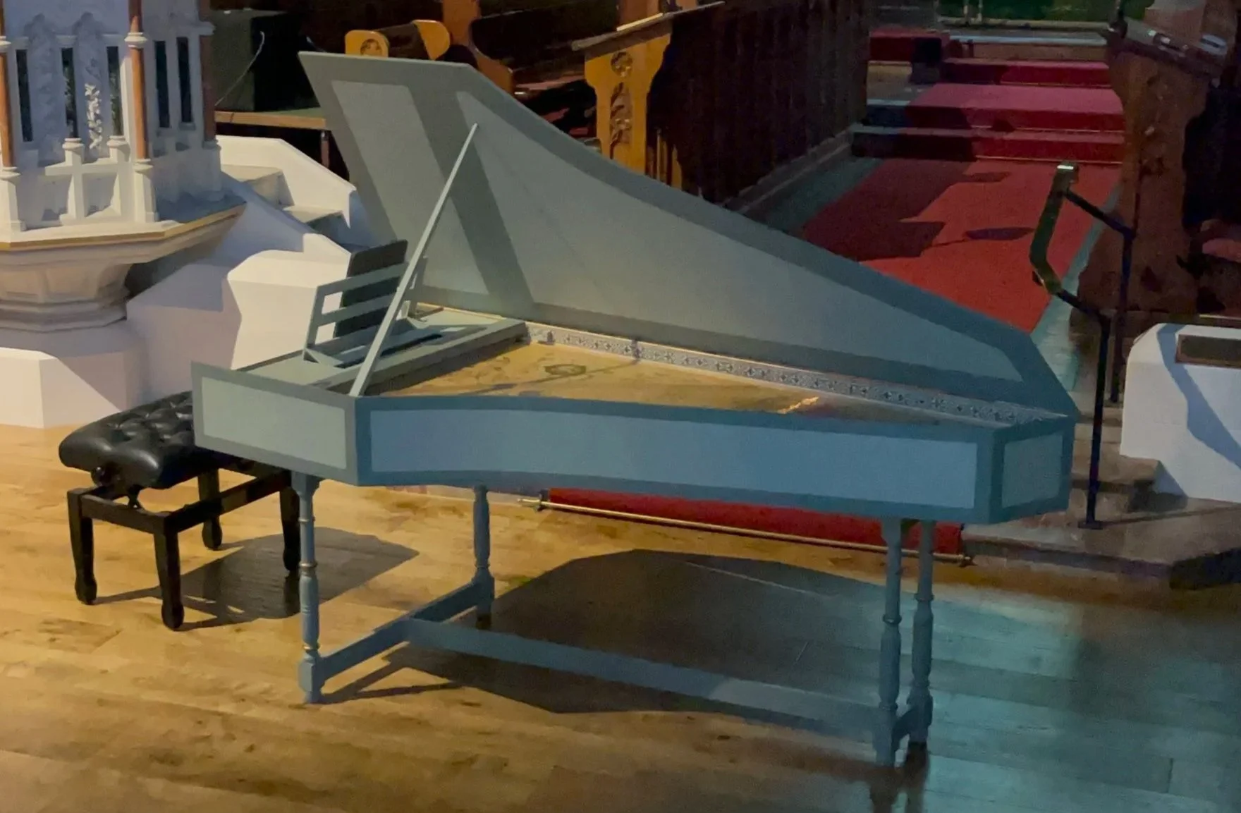 A harpsichord with an open lid on a wooden floor with a black bench nearby in a church. In the background, there are white decorative objects and steps with a red carpet.