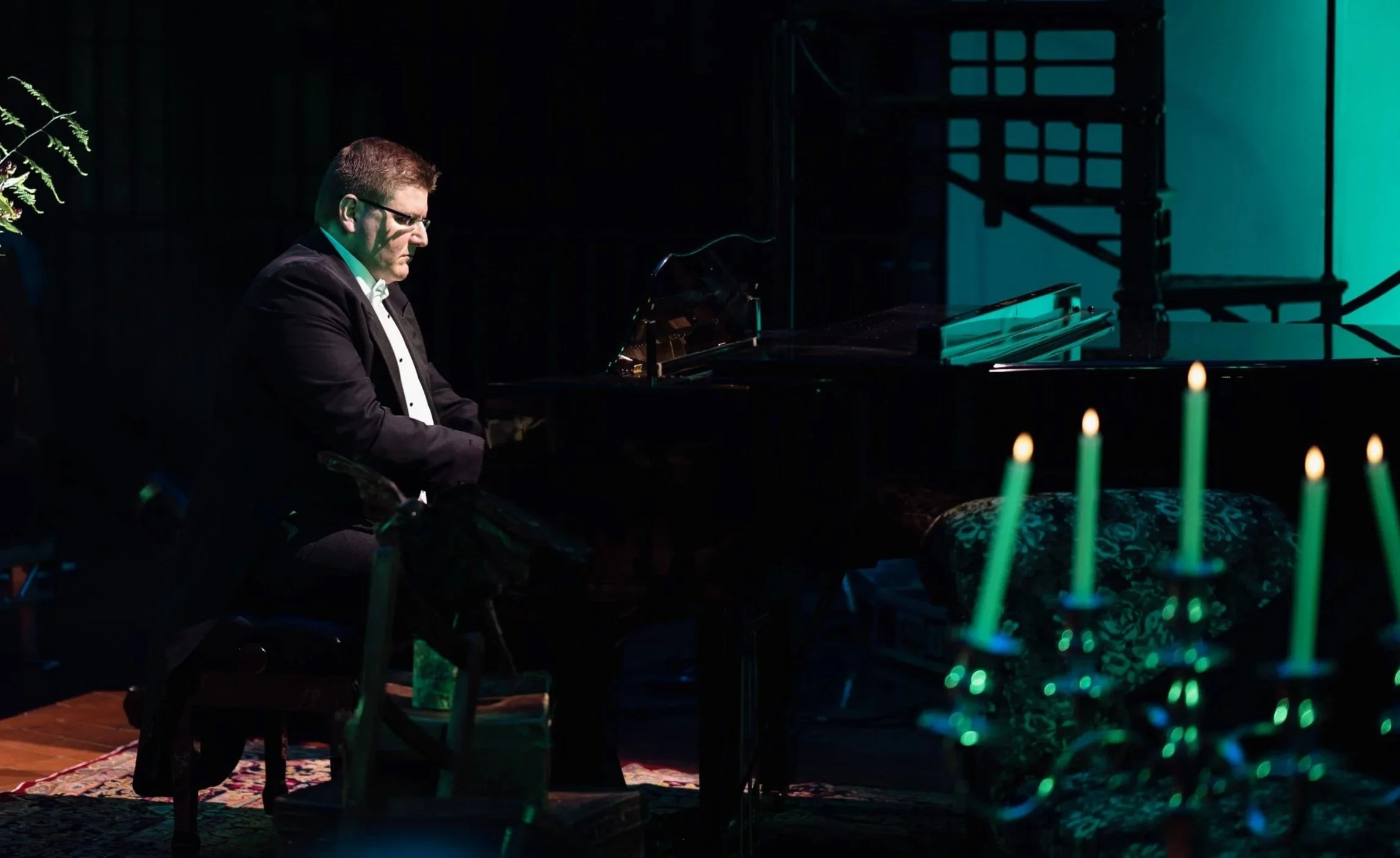 A man in a tuxedo playing the piano in a dimly lit room with green candles in the foreground.