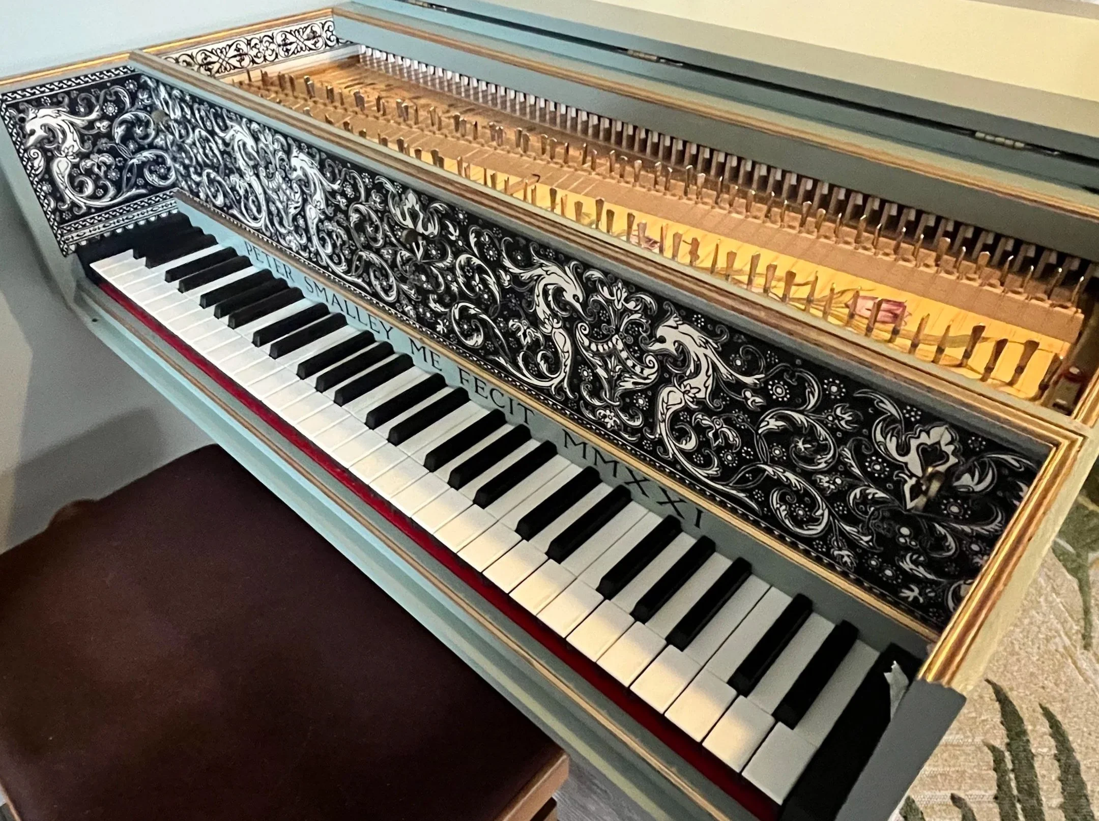 Decorative harpsichord keyboard with ornate black and white floral design on the front, open to reveal the strings inside, with a matching stool nearby.