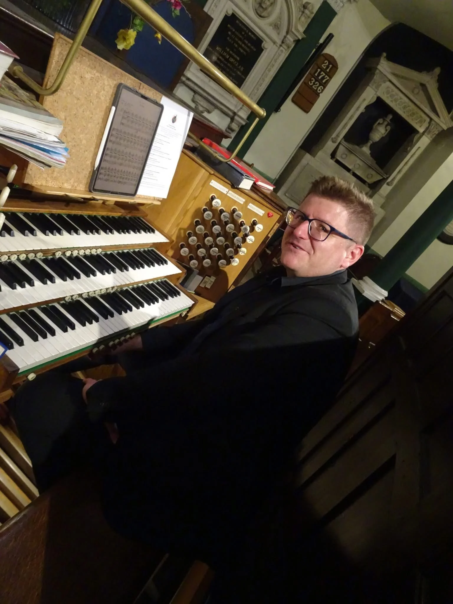 A man with glasses sitting at an organ in a church or historic building, surrounded by church furnishings and artwork.
