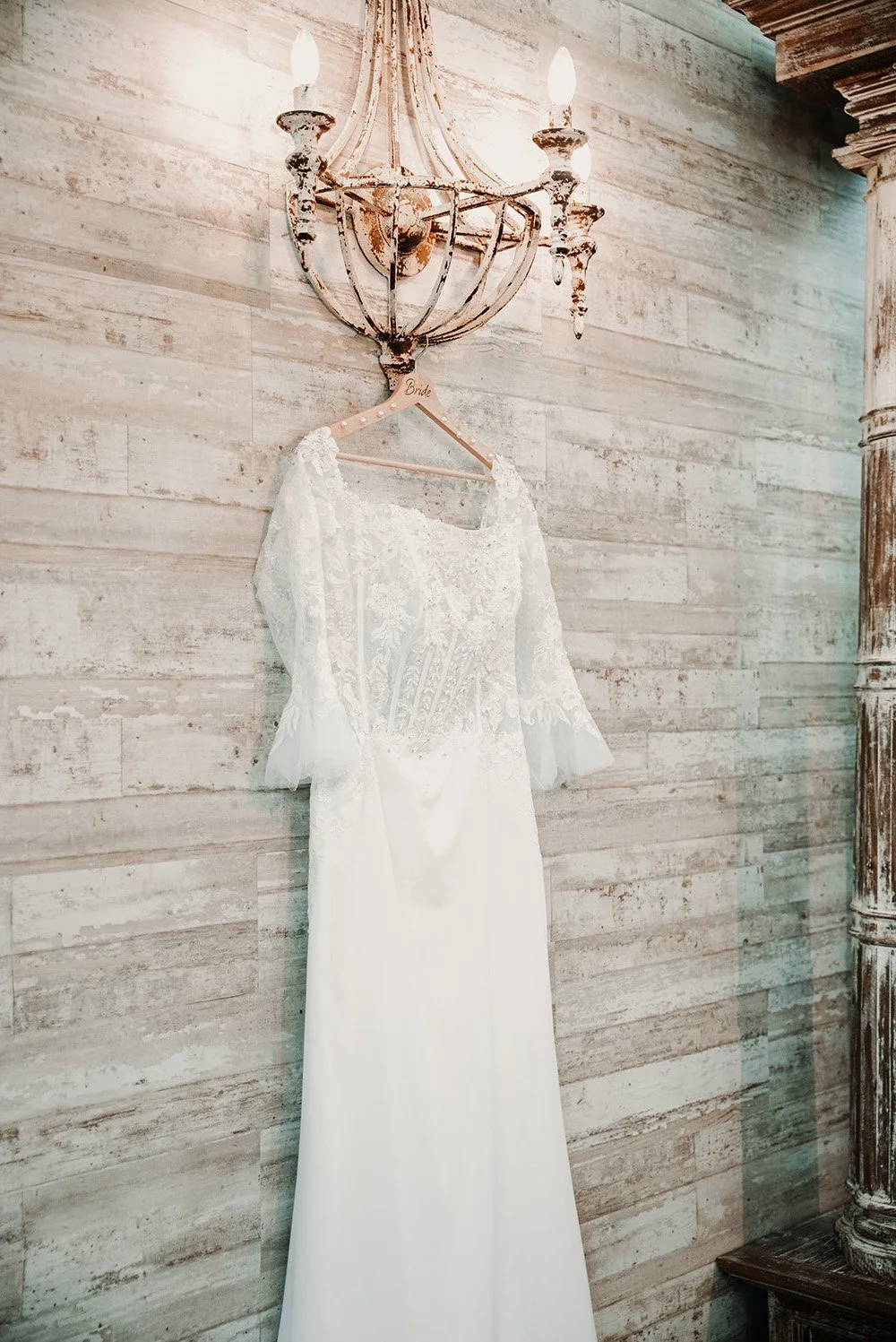 A white wedding dress hanging on a wooden hanger labeled 'Bride' on a rustic wall, illuminated by a vintage chandelier with exposed light bulbs.