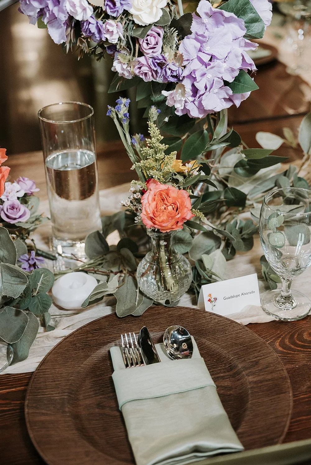 A beautifully set dining table with a flower arrangement, water glass, wine glass, and place setting with a wooden charger and wrapped utensils.