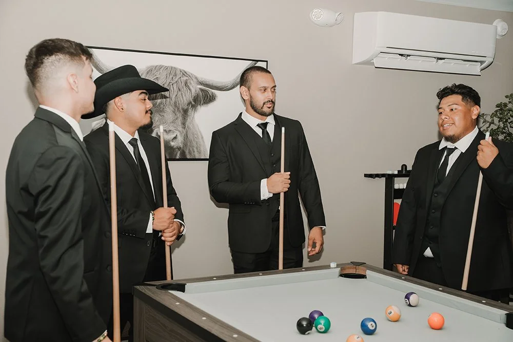 Five men in suits playing pool. One man appears to be explaining or giving instructions to the others. The room has a painting of a buffalo or bison and an air conditioning unit.