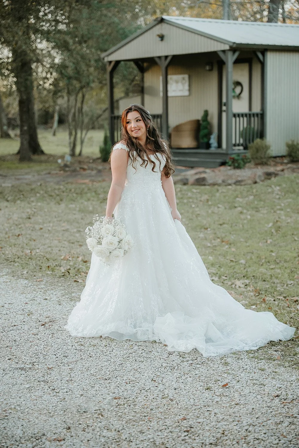 Bride in a white wedding gown holding a bouquet of white roses, standing outdoors near a small gray building with a porch, surrounded by trees and grass.