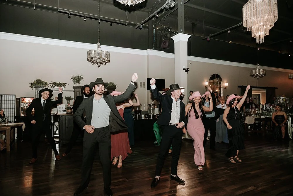 A group of people dressed in Western and cowboy-themed attire, dancing and celebrating at an indoor event with chandeliers and a festive atmosphere.