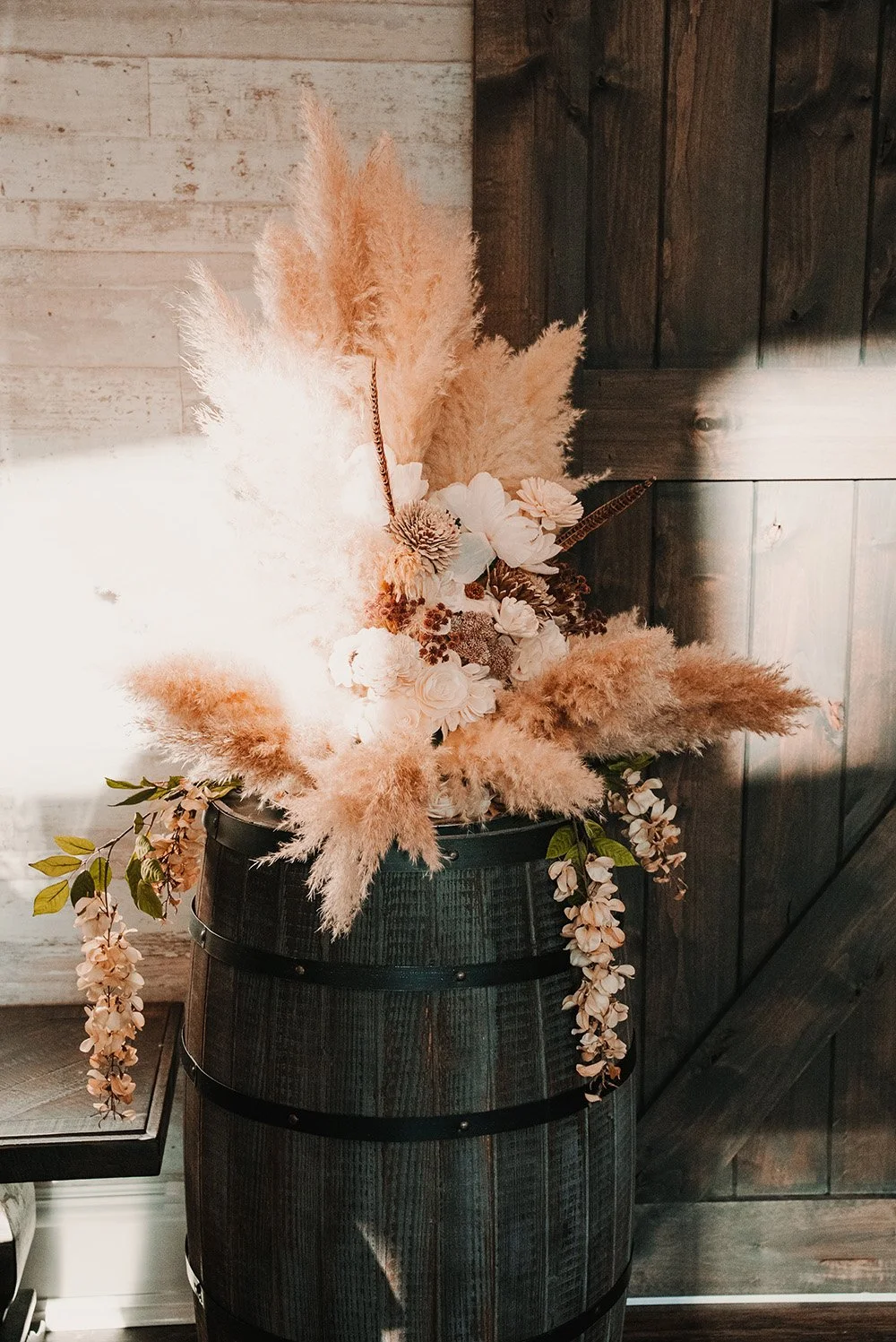 Arrangement of pink and white flowers, including pampas grass, in a black barrel-shaped container against a rustic wooden wall.