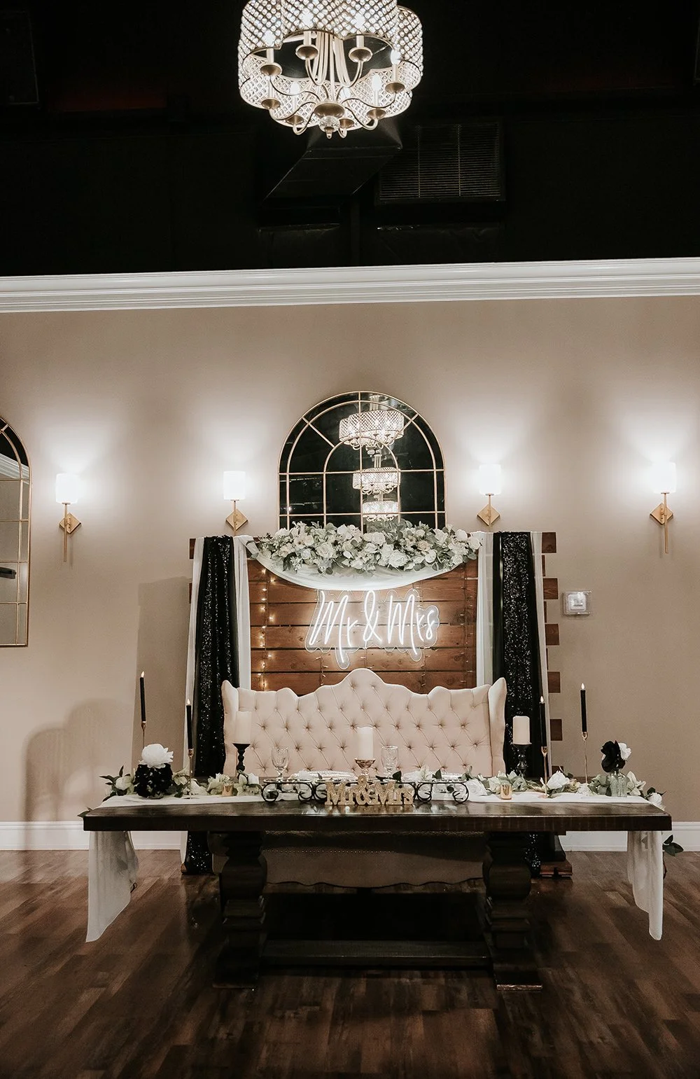 Wedding reception setup with a plush pink tufted sofa, floral arrangements, black candles, and a neon sign that says 'Mr. & Mrs.' The background features a wooden wall, a decorative mirror, and a floral arrangement on top of the wall decor.