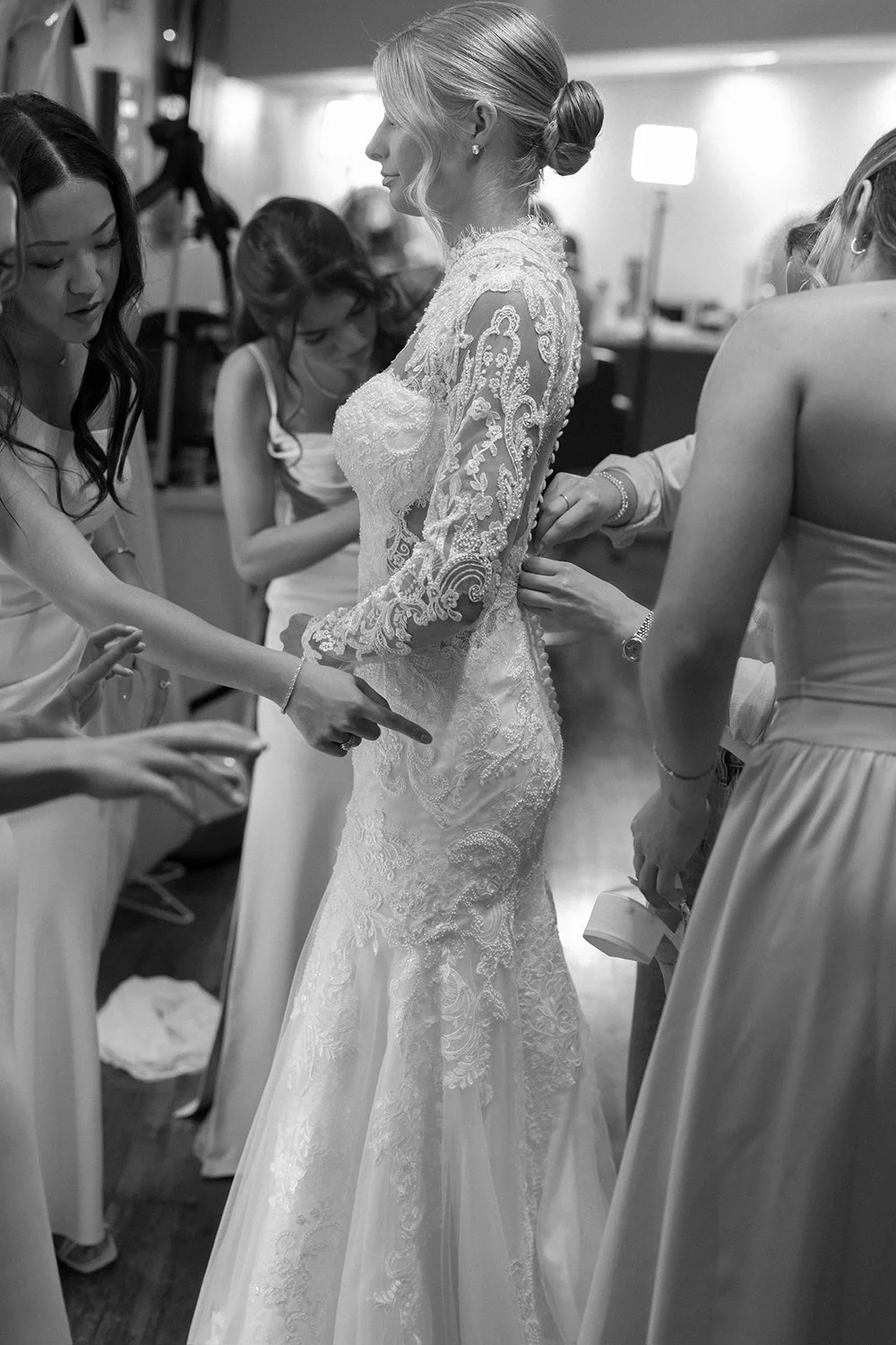 A bride in a lace wedding dress being helped into her gown by her bridesmaids in a dressing room.