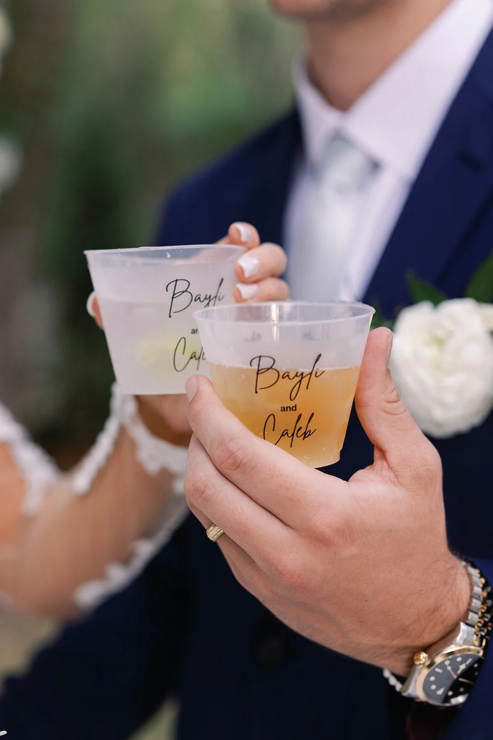 A bride and groom holding plastic cups with drinks to make a toast, one with a pale yellow beverage and the other with a clear or light-colored beverage, both cups inscribed with 'Bayli and Caleb'.