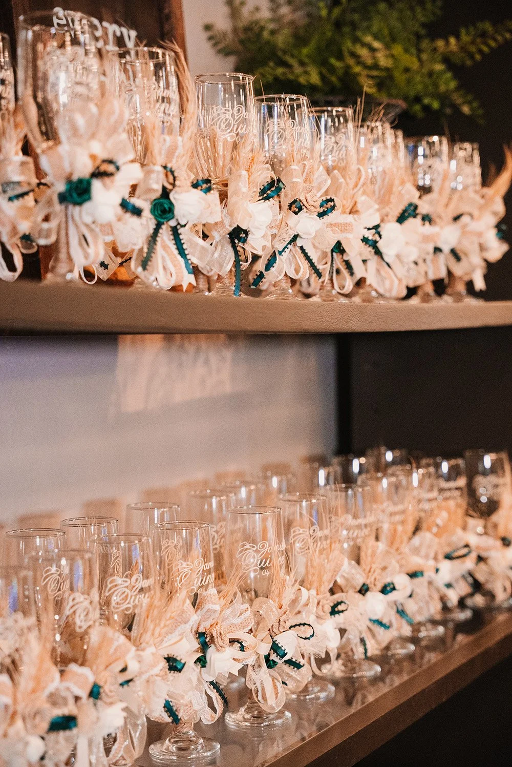 A display of decorated glasses with ribbon bows, small bouquets, and embroidered text, arranged on a shelf at a celebration or event.