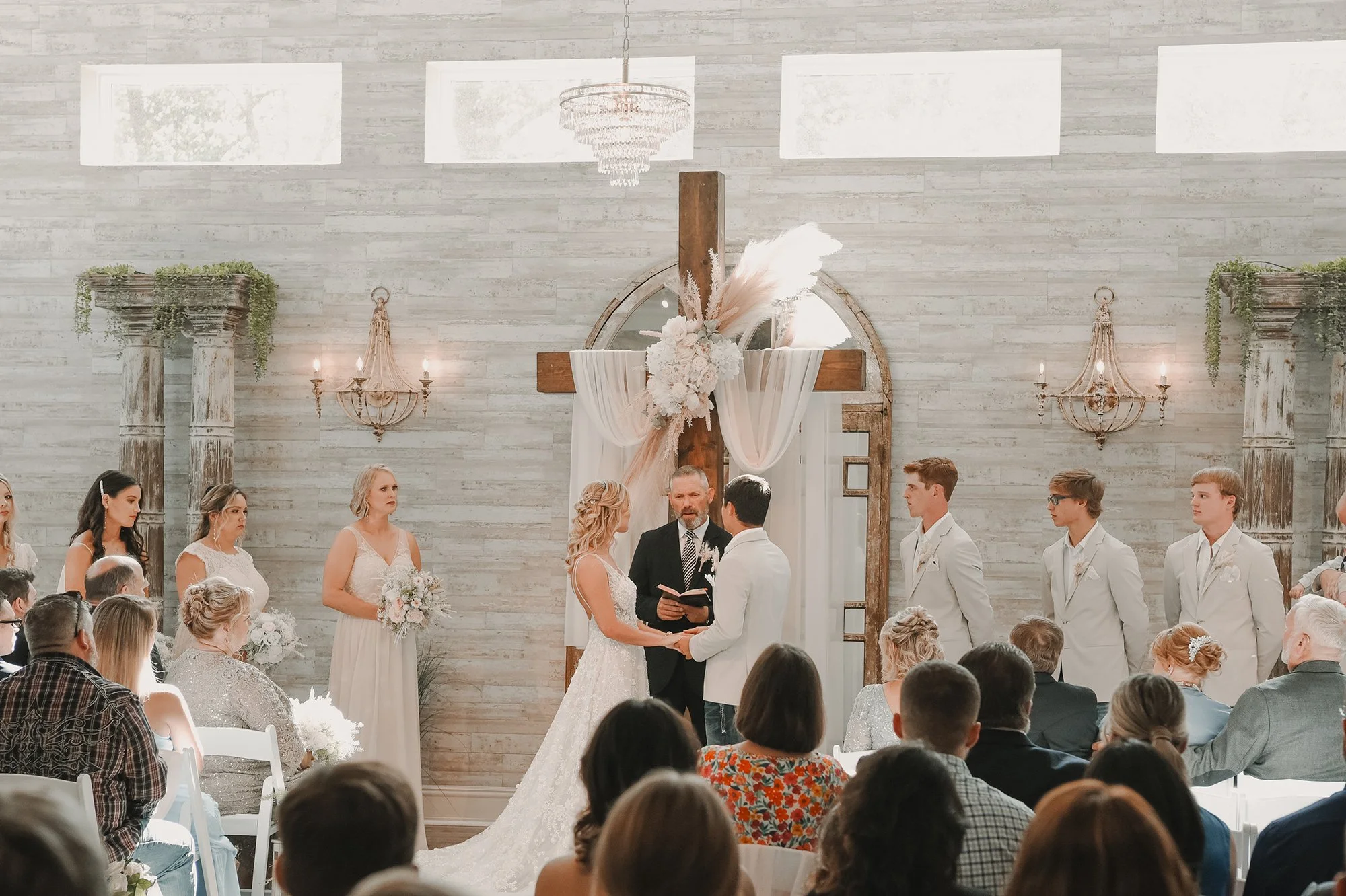 A wedding ceremony inside a church with a bride and groom exchanging vows, officiated by a person holding a book. The church has a rustic modern decor with a large cross decorated with flowers and draped fabric as the background. Guests seated in chairs are watching. The ceremony is surrounded by elegant wall sconces and floral columns.