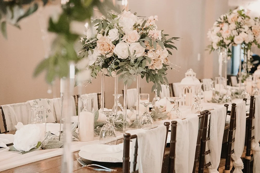 Elegant banquet table decorated with tall floral centerpieces, candles, and glassware, in a softly lit room.