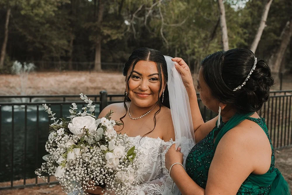 A bride holding a bouquet of white flowers with a woman adjusting her veil outdoors by a pond or lake with trees in the background.