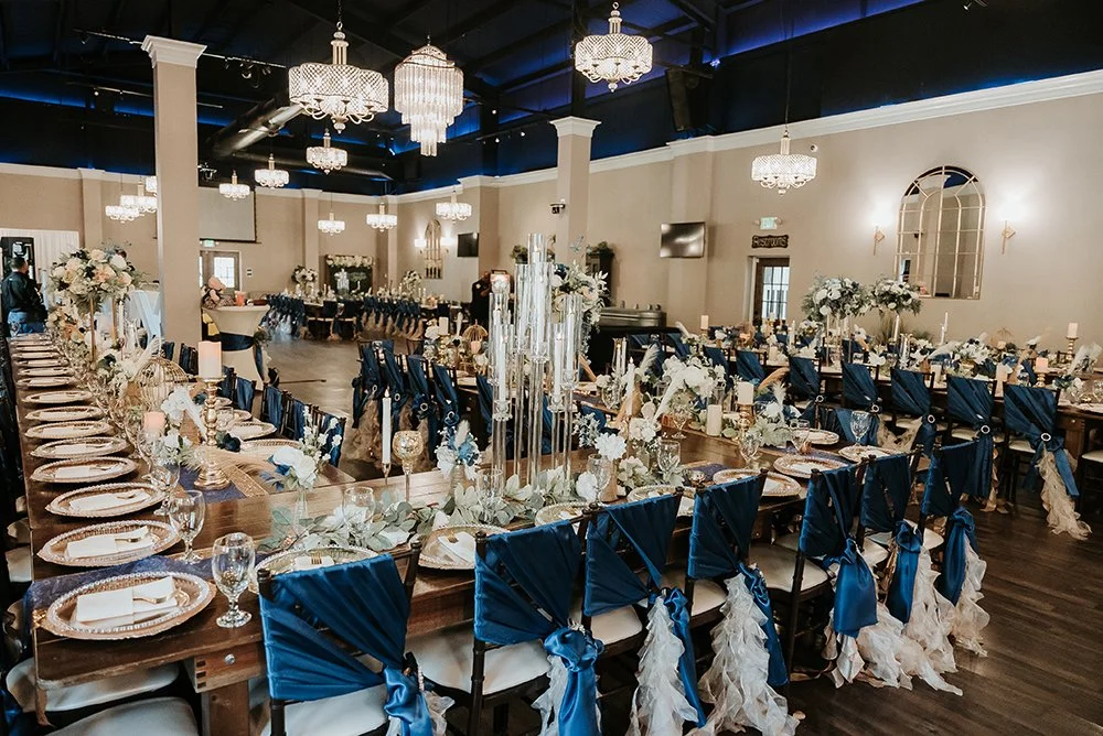 Elegant banquet hall decorated for a wedding reception with long wooden tables, navy blue chair sashes, floral centerpieces, and chandeliers.