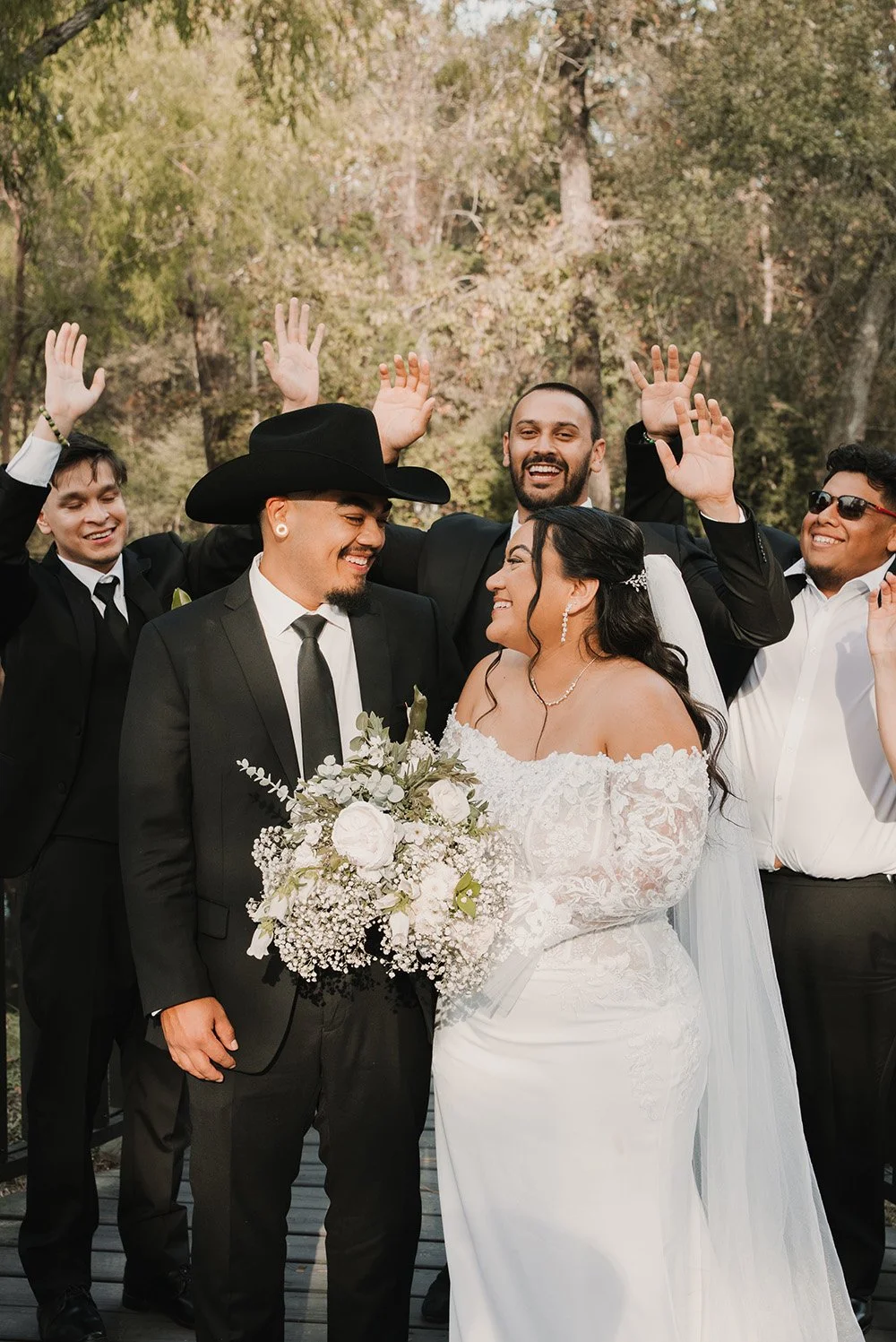 Bride and groom with wedding party outdoors, smiling, bride holding a bouquet, groom in a black suit and cowboy hat, friends celebrating in background.