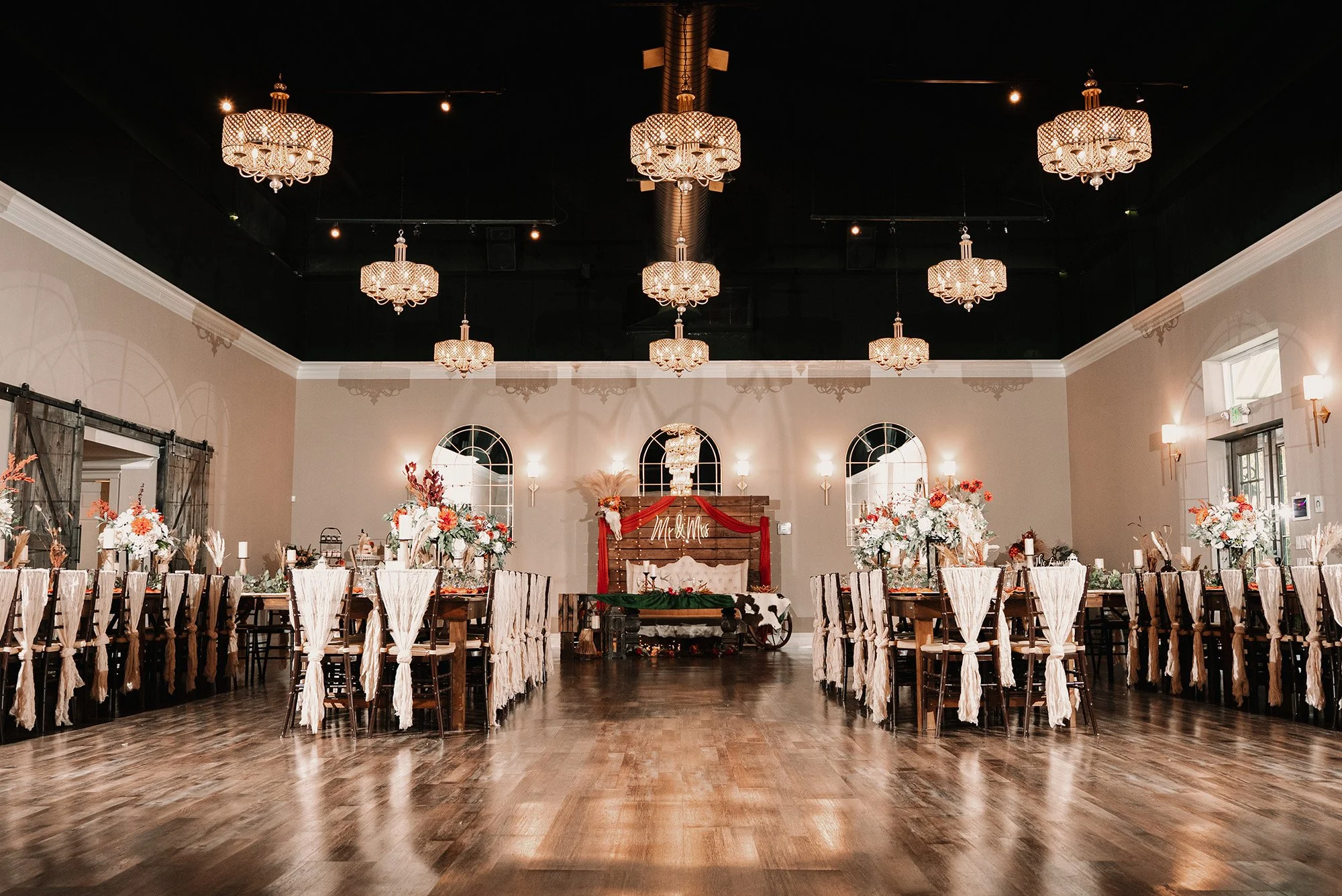 A decorated banquet hall with long tables, floral centerpieces, and a wooden backdrop with a red fabric drape reading "Mr & Mrs". The ceiling has ornate chandeliers and pendant lights, with windows on the side walls.