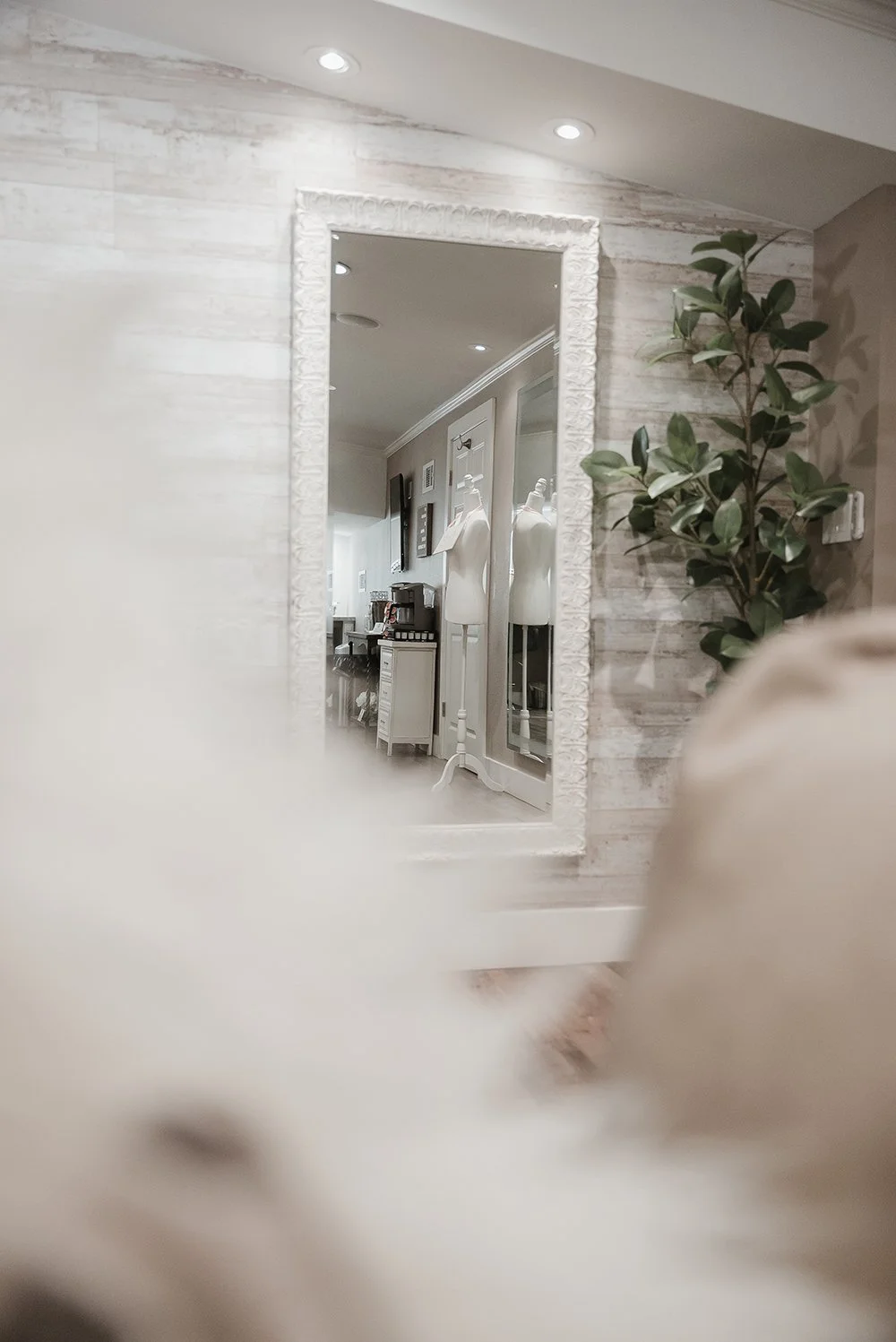 A mirror reflecting a room with mannequins and a shelving unit, decorated with plants and framed pictures, in a cozy, well-lit interior space.