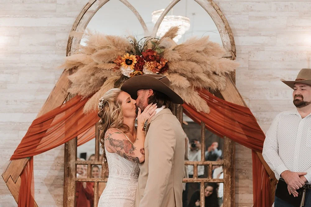 A couple sharing a kiss during their wedding ceremony, with a man in a cowboy hat on the side, under a rustic floral and pampas grass decoration.