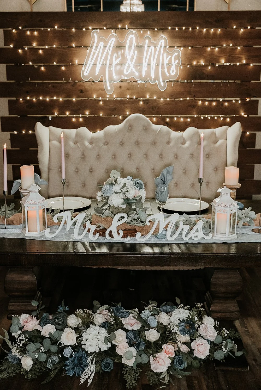 Decorated wedding reception table with a beige tufted high-back sofa, candles, lanterns, floral arrangements, and a neon sign that reads 'Mr. & Mrs.' in front of a wooden plank wall with string lights and a neon sign that reads 'Mr. & Mrs.'.