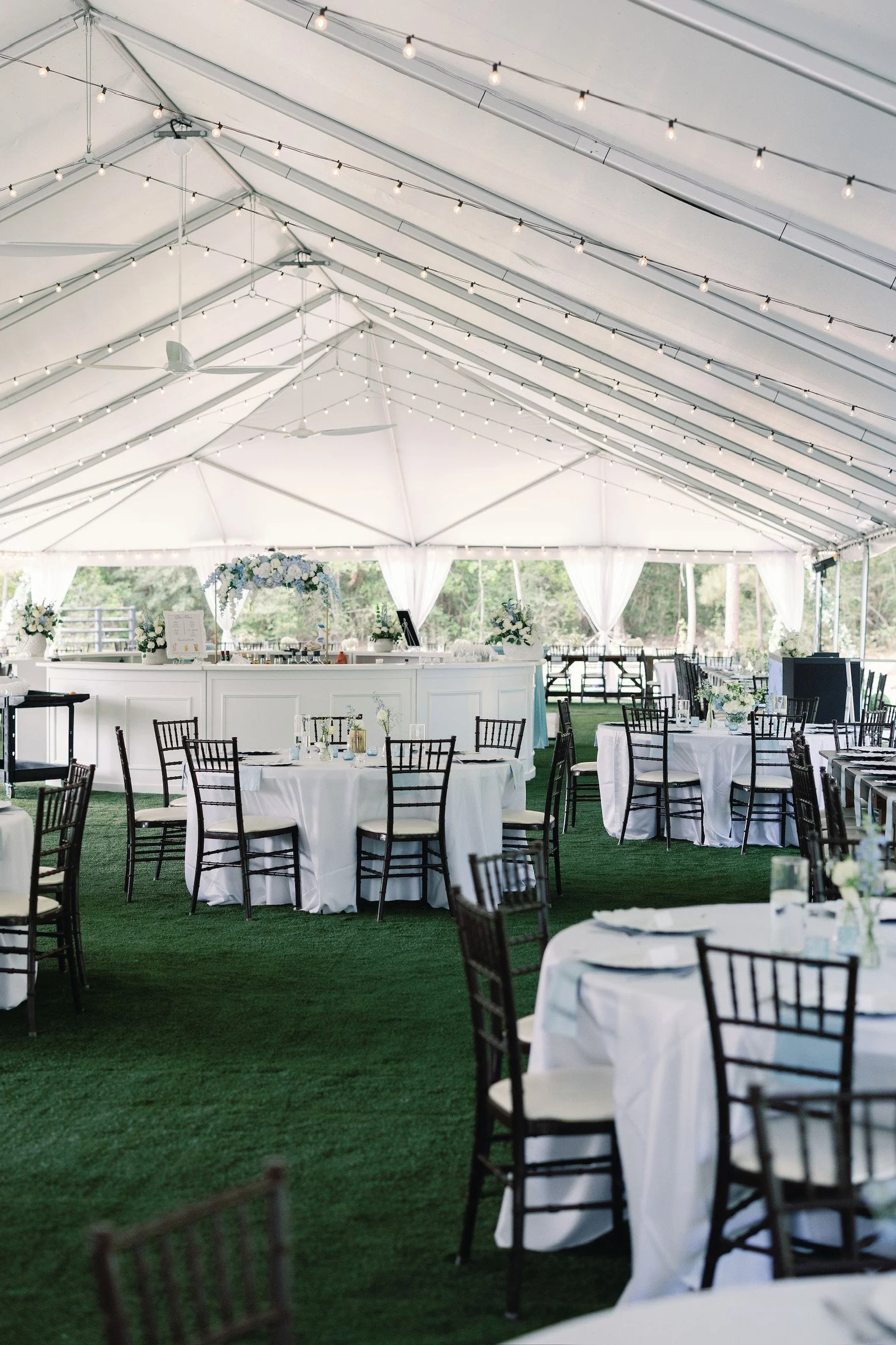 Indoor event tent decorated with string lights, white curtains, tables with white tablecloths, floral centerpieces, and black chairs, set up for a celebration or wedding reception.
