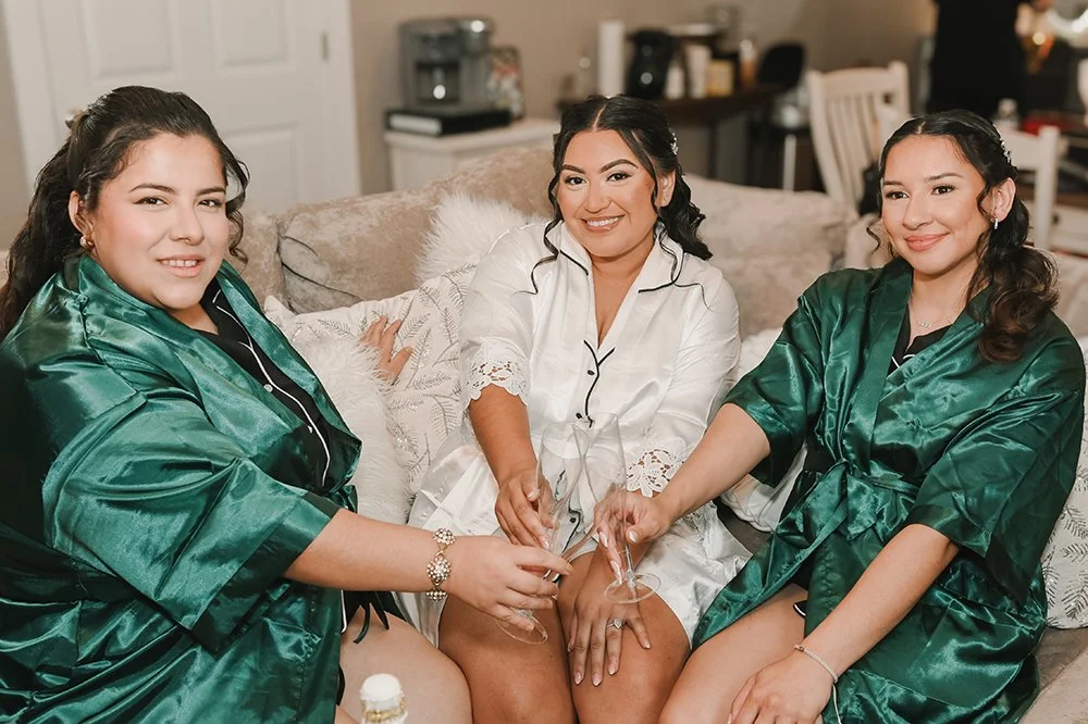 Three women in matching satin pajamas sitting on a couch, holding champagne glasses, smiling at the camera.