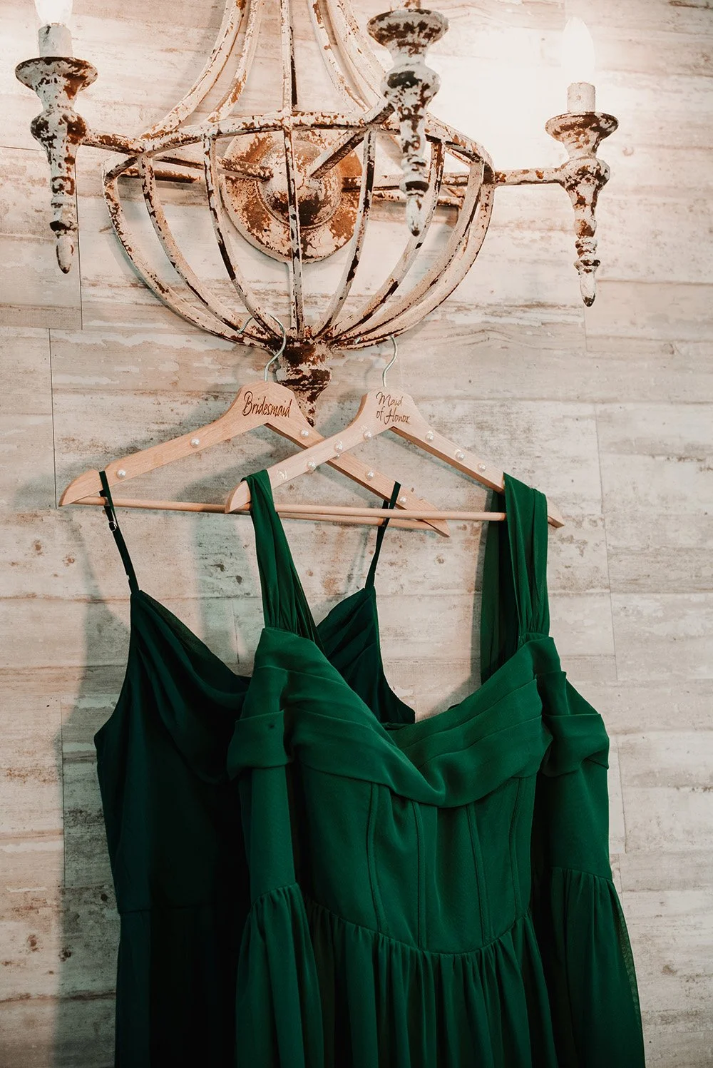 Green bridesmaid dress hanging on a wooden hanger labeled 'Bridesmaid' on a rustic wall with a vintage chandelier