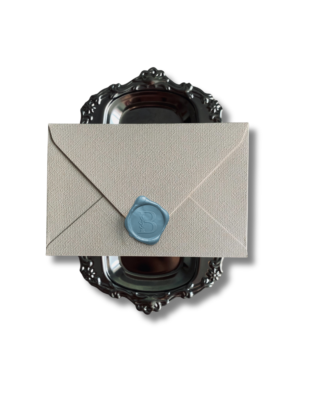 A beige envelope with a blue wax seal labeled 'B', partially covering a decorative silver tray with an ornate border, against a black background.