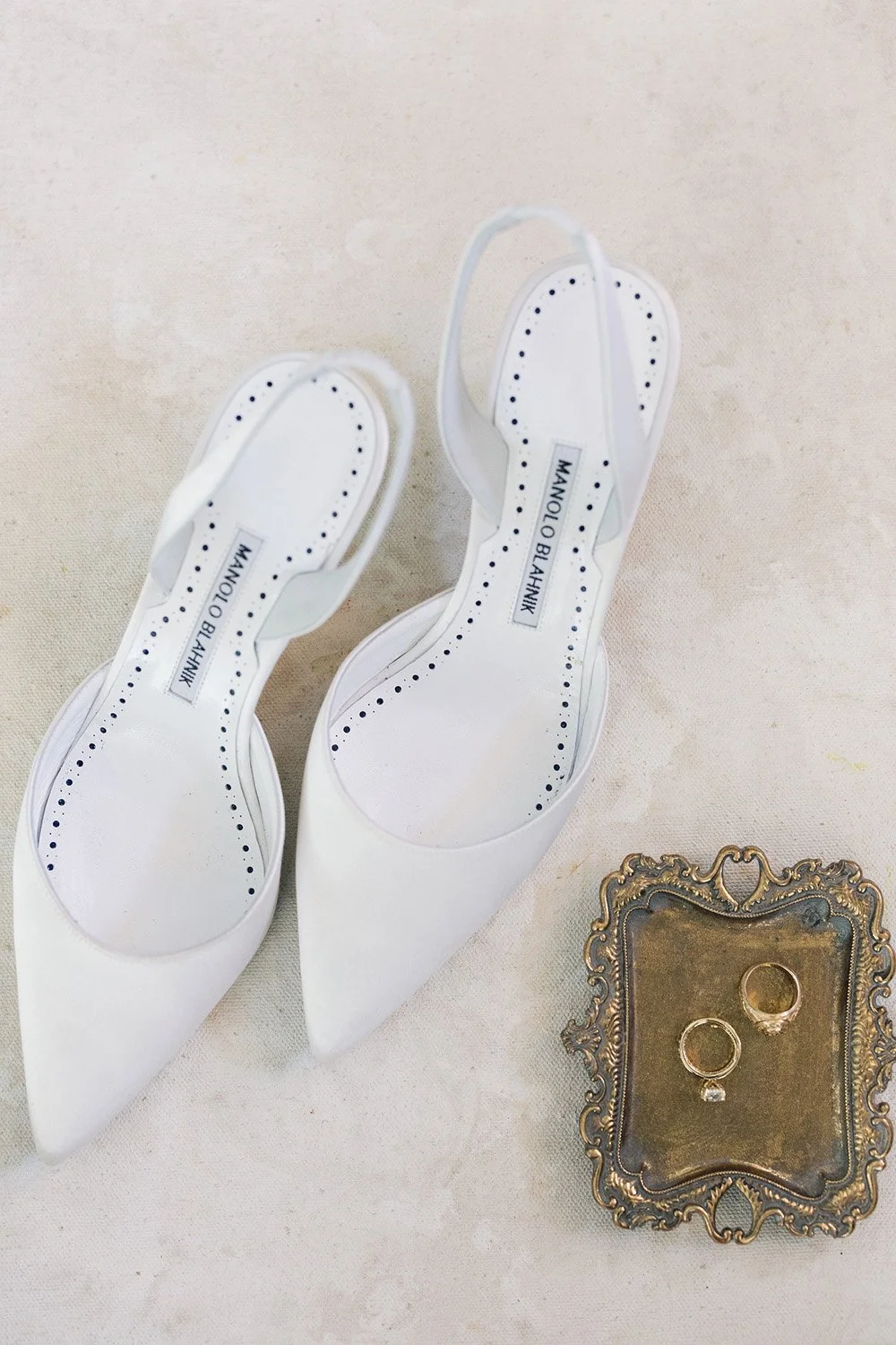 Pair of white Manolo Blahnik high-heeled shoes on a beige surface, with a decorative gold tray holding two rings nearby.