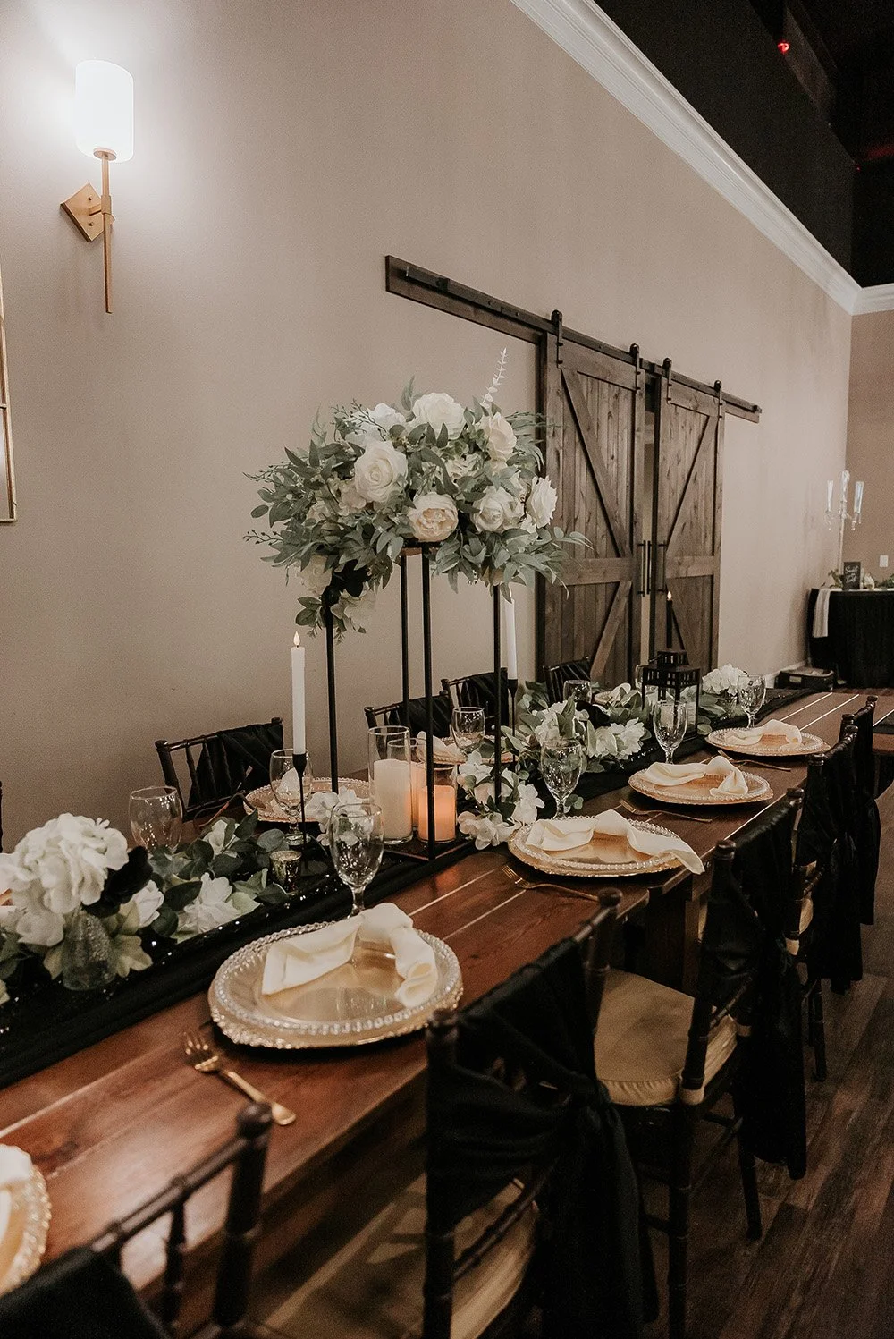 Decorated dining table with floral centerpiece, candles, place settings, and black chairs in a rustic event space.