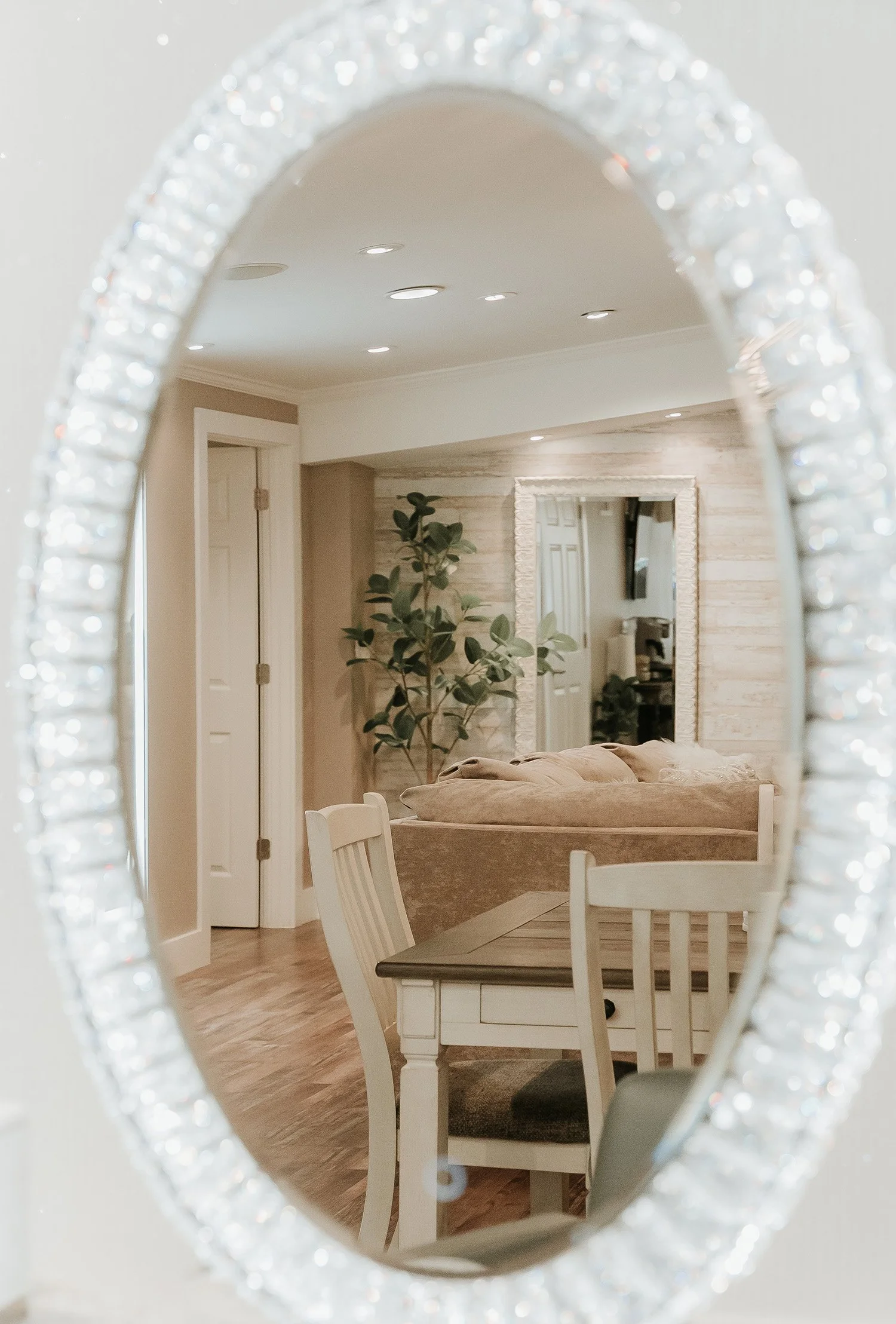 Interior of a cozy living room with a beige sofa, a large mirror on a wall with light-colored wooden panels, a potted plant, and a white door, reflected in a decorative mirror with a beaded frame.