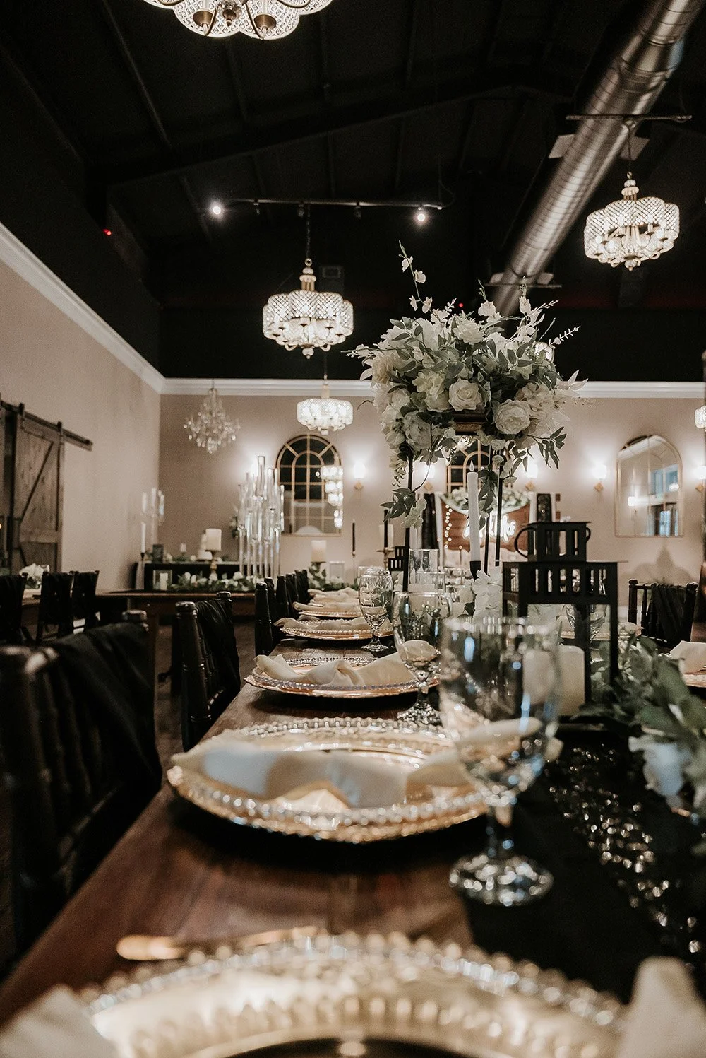 Elegant banquet hall decorated for a formal event with a long wooden table, white floral centerpiece, ornate gold-rimmed plates, and glassware, with chandeliers hanging from the dark ceiling.