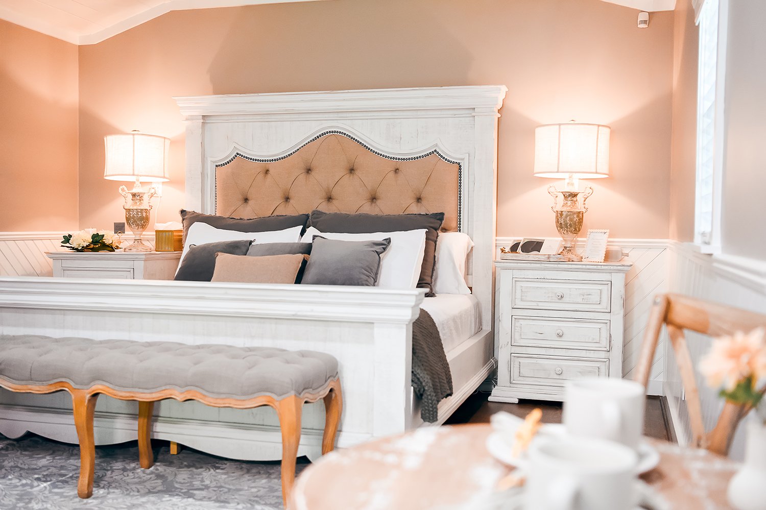 A bedroom with a large white bed, beige tufted headboard, dark gray and beige pillows, bedside tables with lamps, and a dining table with flowers and cups in the foreground.