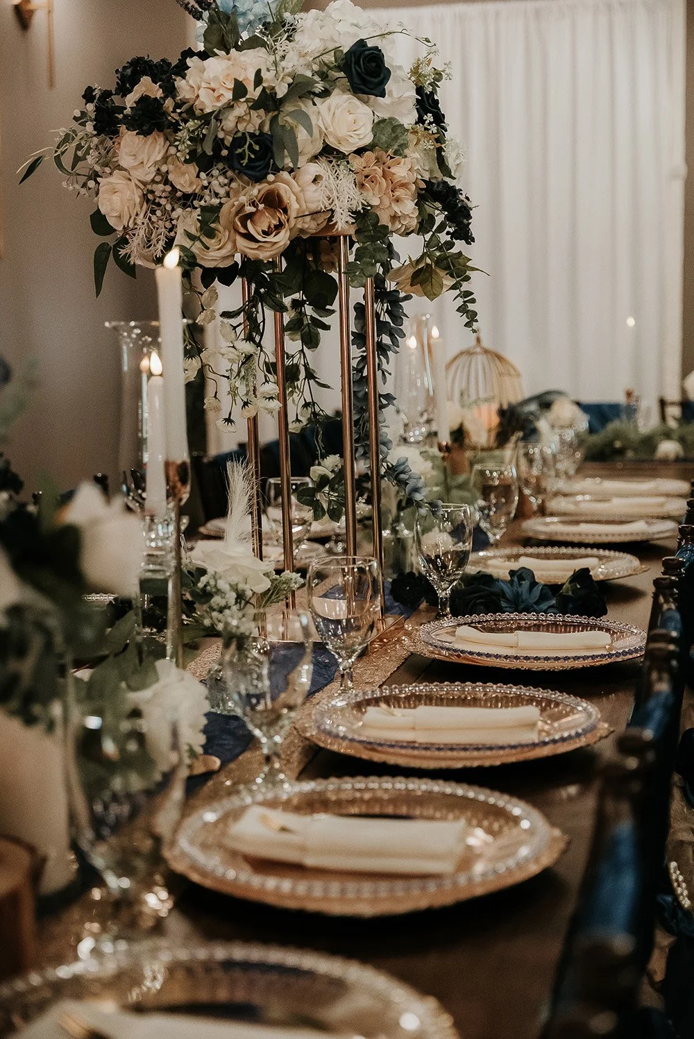 Elegant dining table setting with tall floral centerpiece, candles, and gold-rimmed plates with napkins.