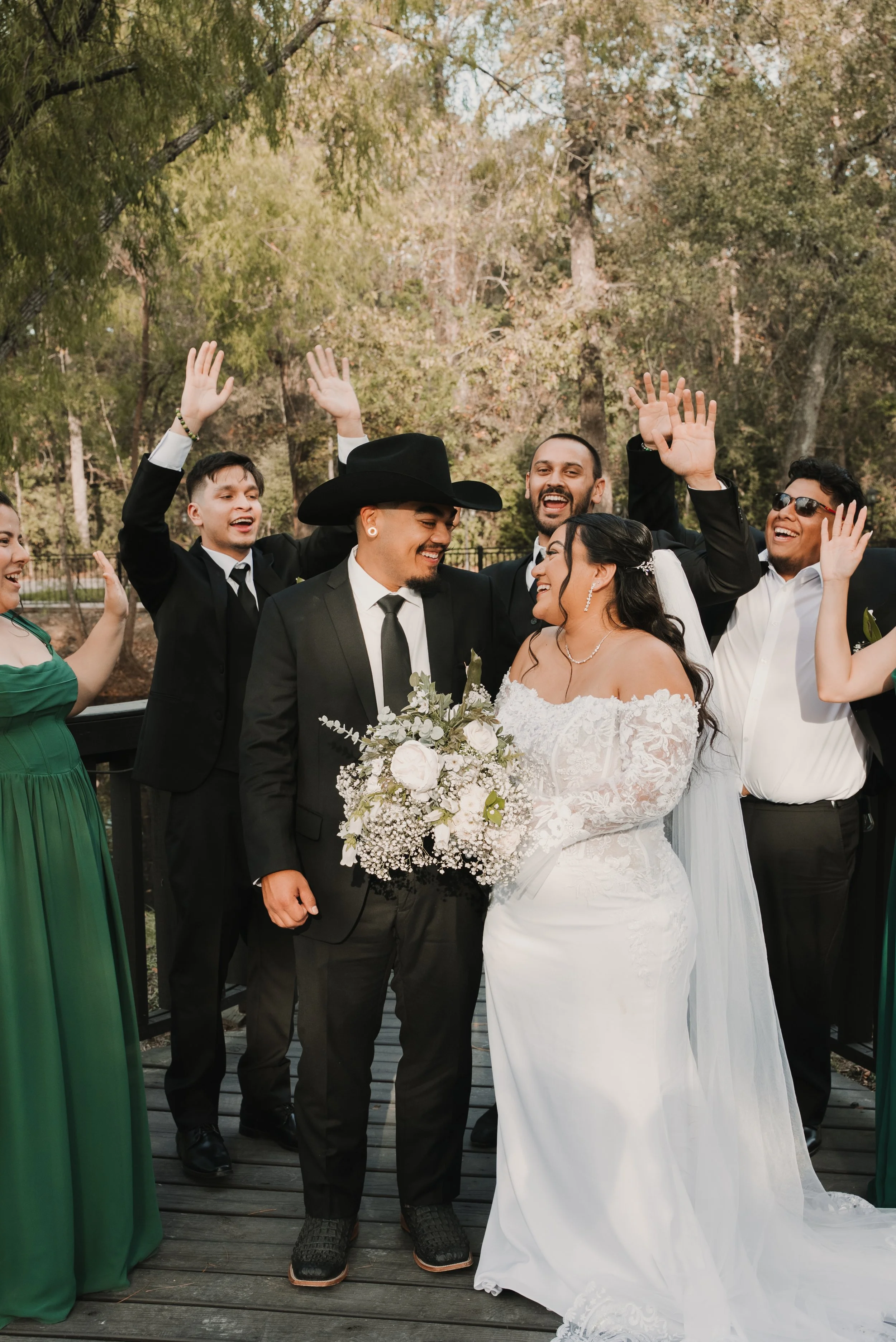 A group of people celebrating at a wedding, with the bride and groom in the center surrounded by friends raising their hands and smiling outdoors in a wooded area.