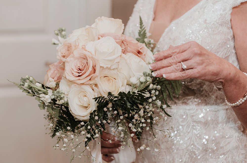 A person in a white, sparkly wedding dress holding a bouquet of light pink and white roses with greenery and small white flowers.