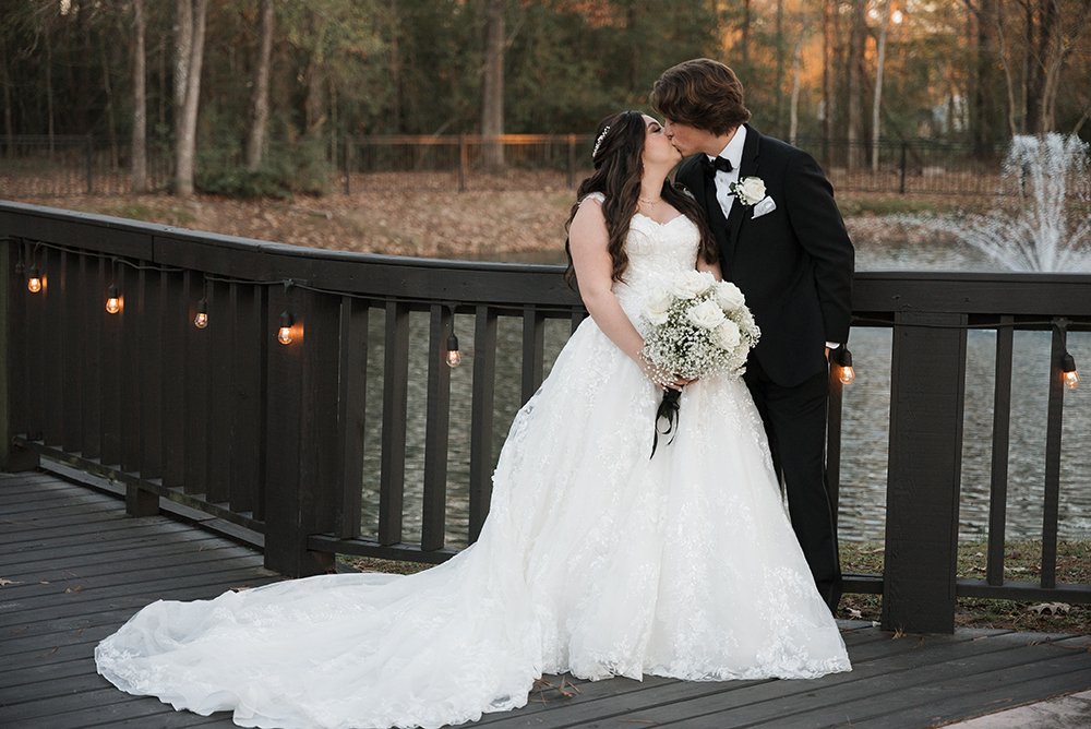 A bride and groom sharing a kiss on a wooden bridge overlooking a pond with a fountain, surrounded by trees during sunset. The bride is wearing a white wedding gown and holding a bouquet, and the groom is in a black tuxedo.