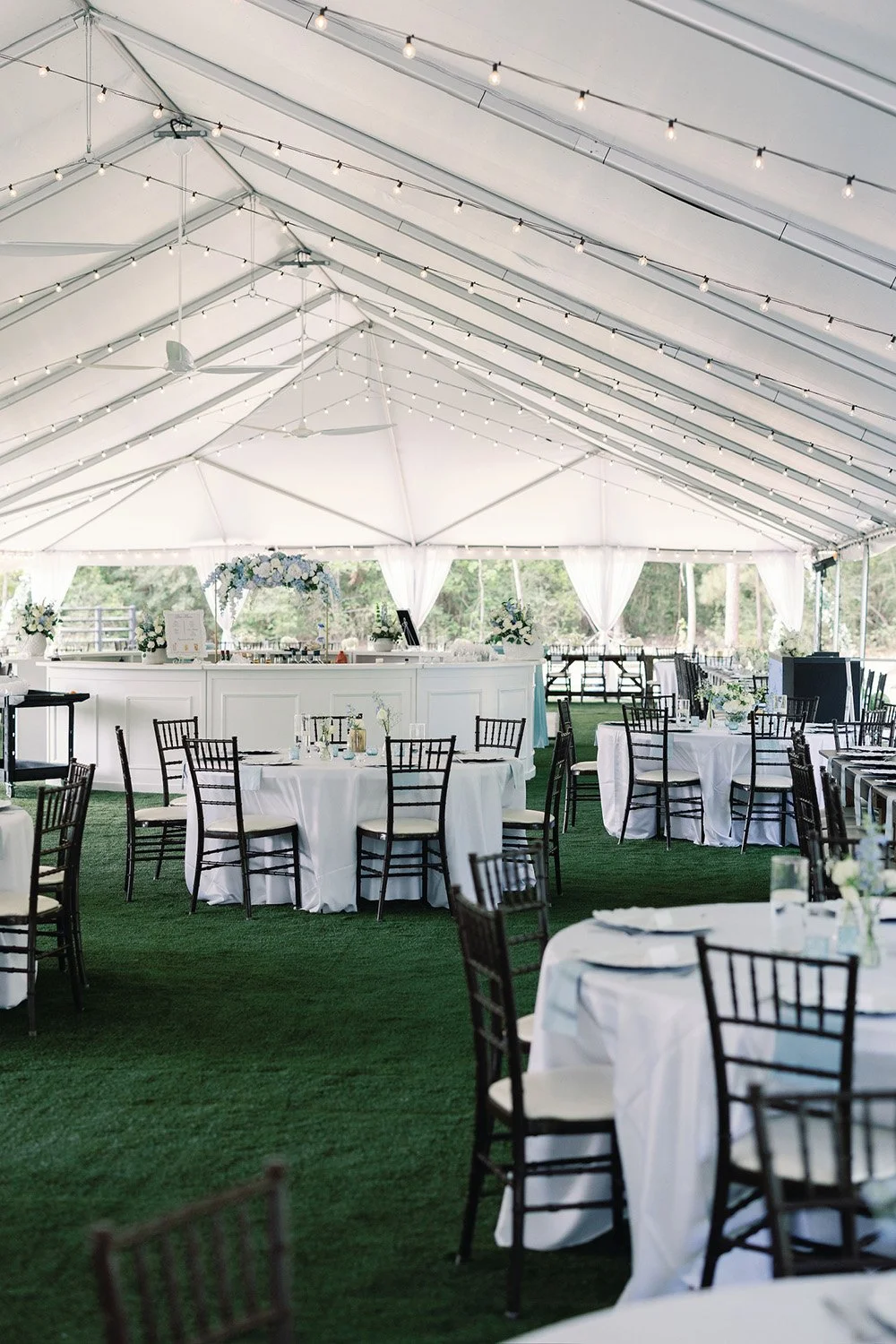 Elegant outdoor tent wedding reception with round tables, white tablecloths, black chairs, floral centerpieces, string lights, and a bar area at the back.